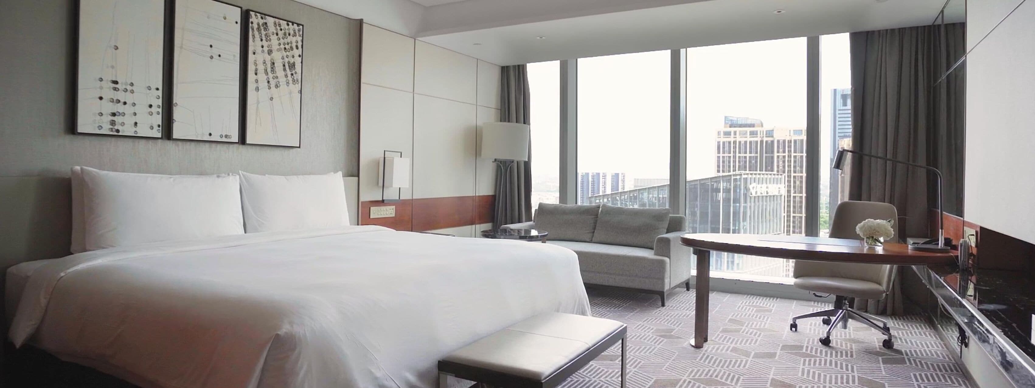 A hotel room with a large bed, a desk, a chair, and a view of the city through large windows.