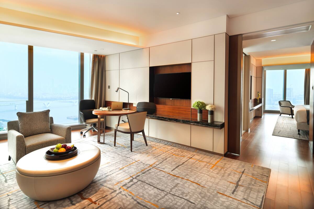 A modern hotel room with a large window, a flat-screen TV, a dining area, and a seating area with a fruit bowl.