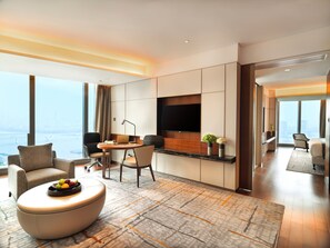 A modern hotel room with a large window, a flat-screen TV, a dining area, and a seating area with a fruit bowl.