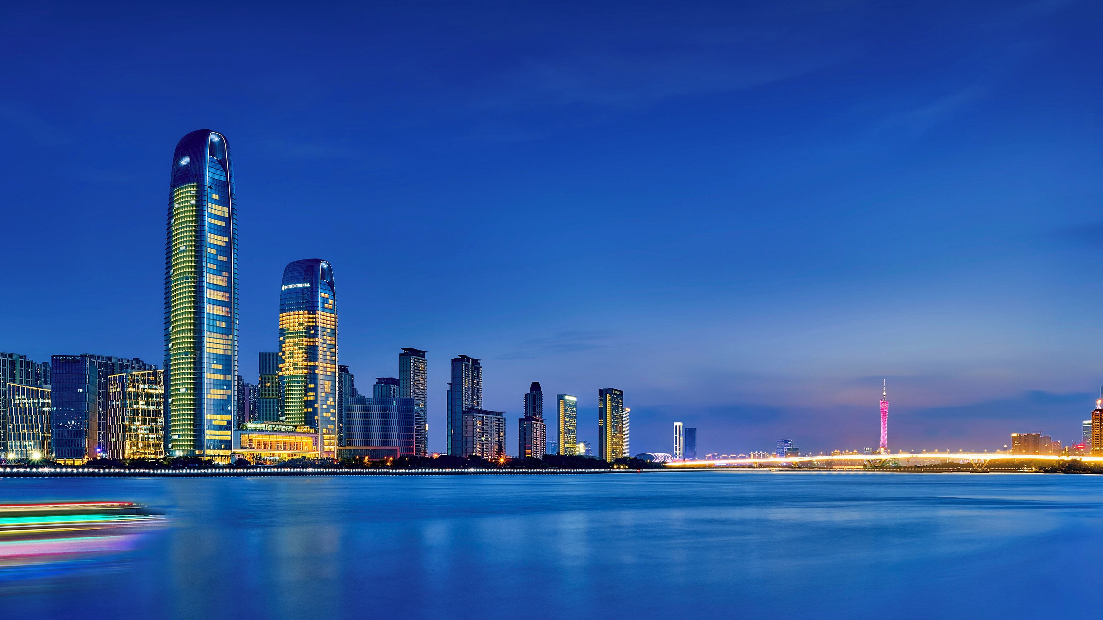 A city skyline with modern high-rise buildings, a calm waterfront, and a bridge in the distance.