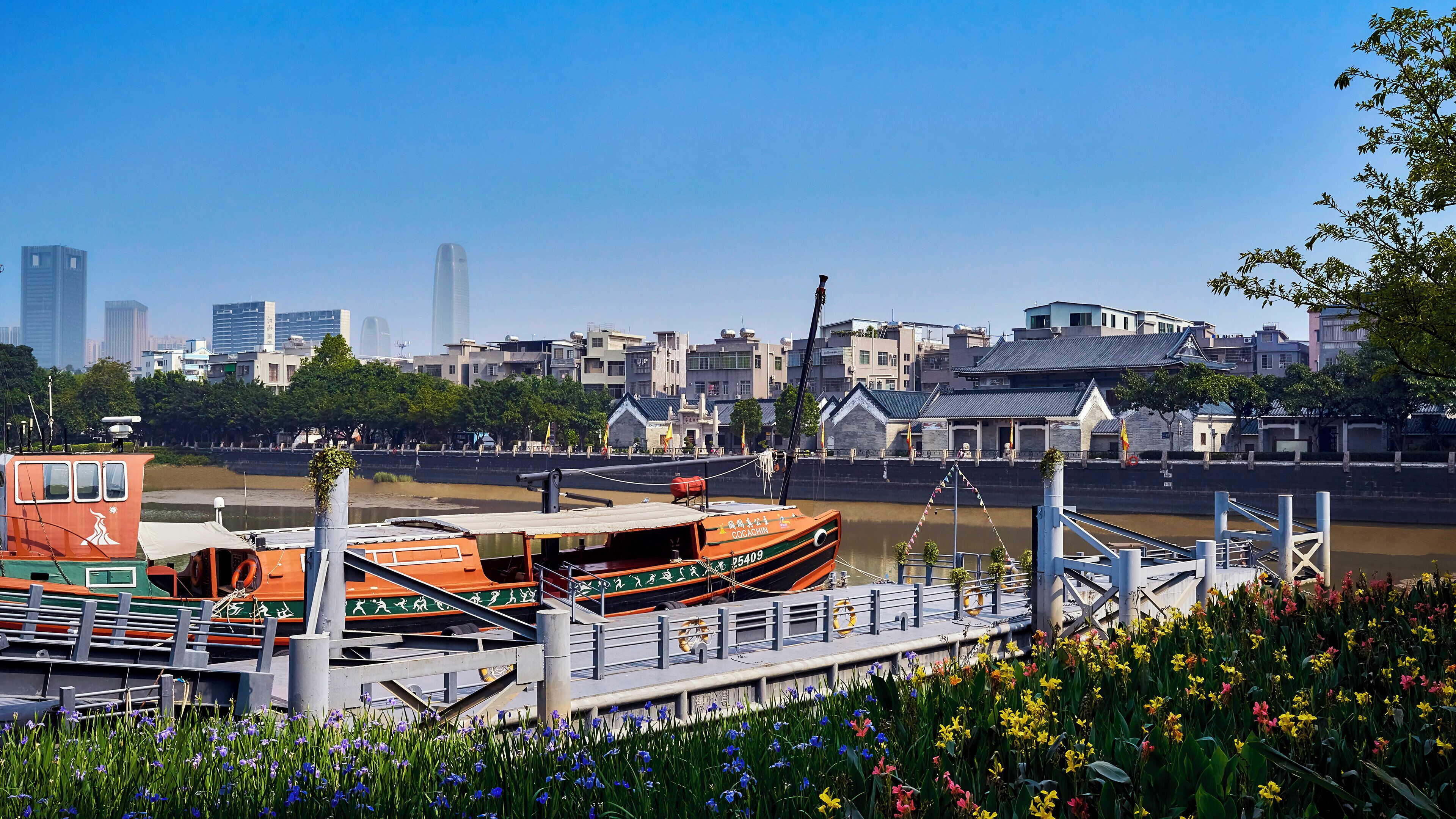A riverfront with a traditional boat docked, a bridge, and a cityscape with modern buildings in the background.
