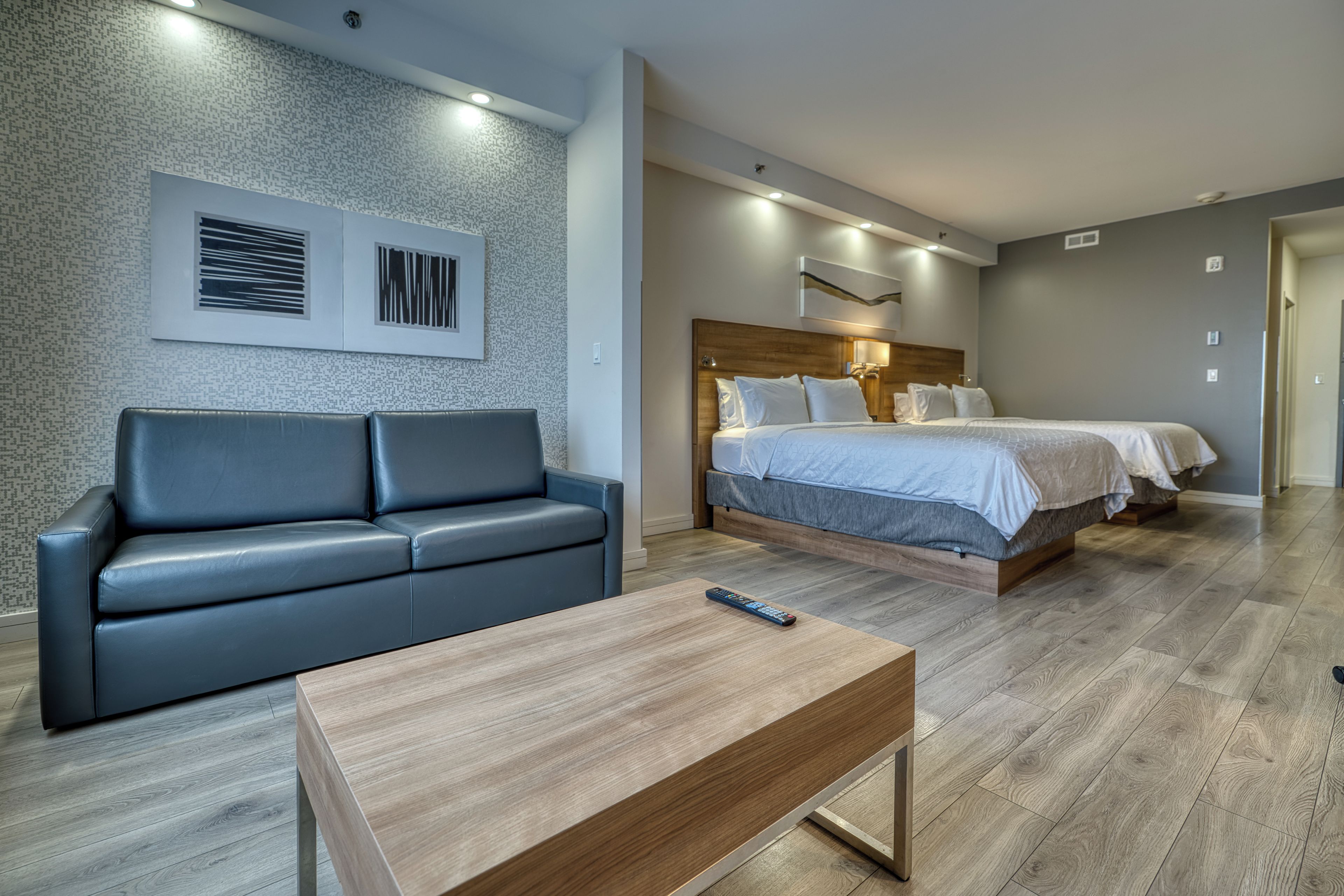 A modern hotel room with a grey sofa, a wooden coffee table, and a double bed with a wooden headboard.