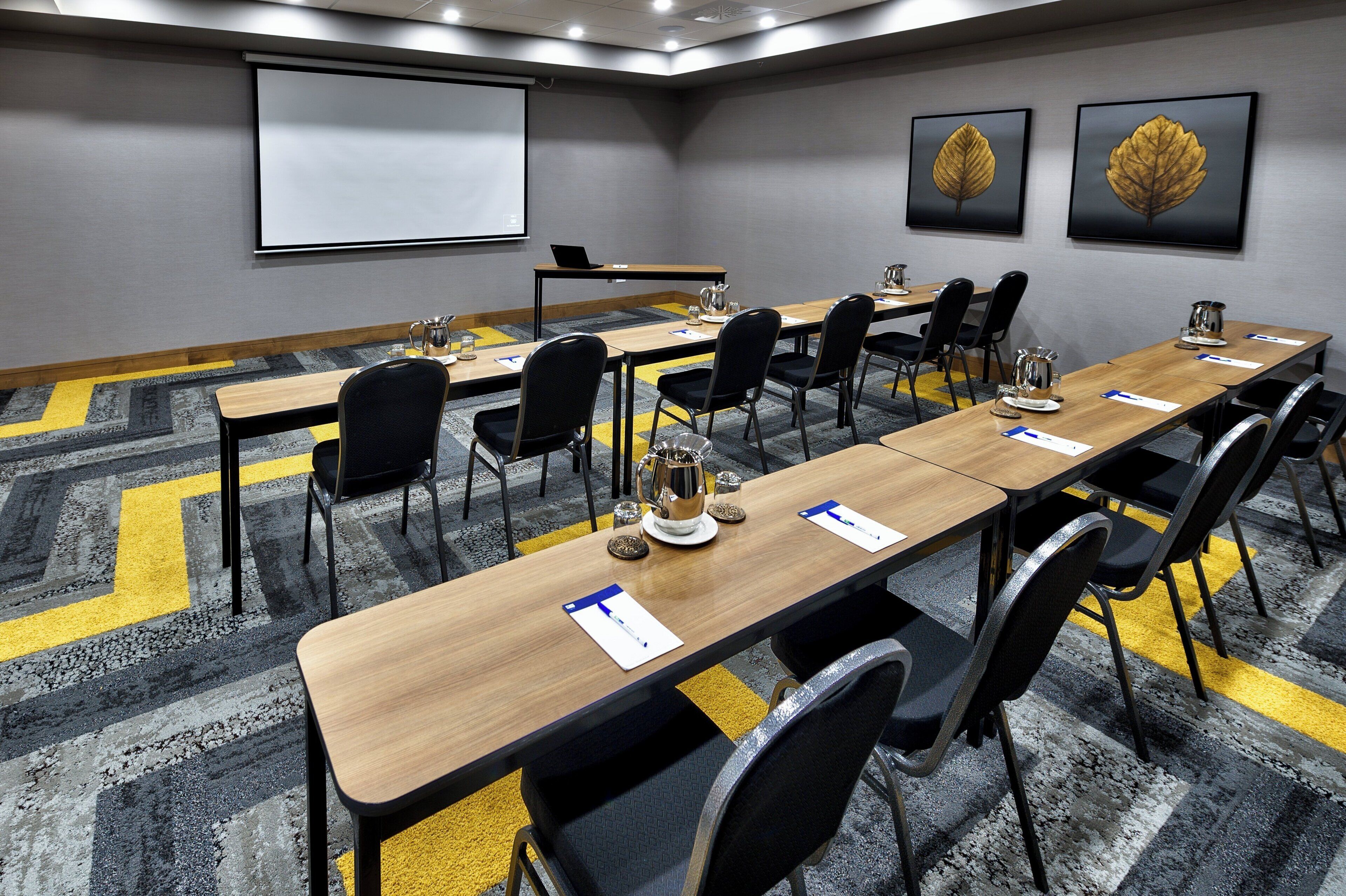 A conference room with a long table, chairs, a projector screen, and two framed leaves on the wall.