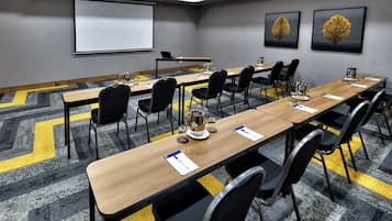 Uma sala de conferência com uma mesa comprida, cadeiras, uma tela de projetor e duas folhagens emolduradas na parede.