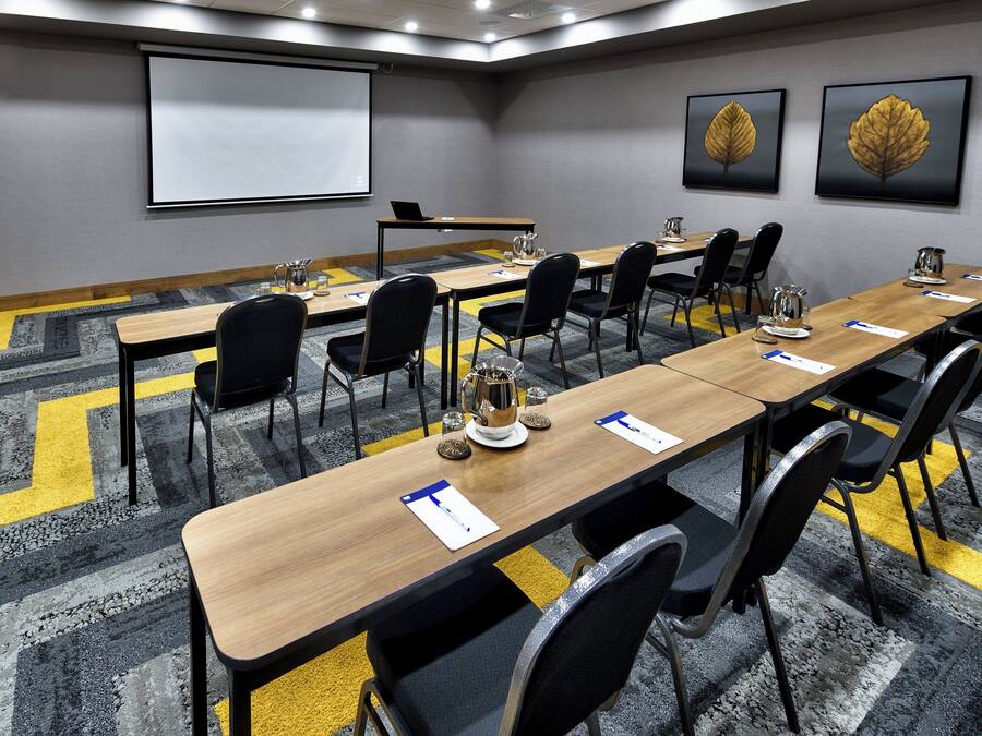 Uma sala de conferência com uma mesa comprida, cadeiras, uma tela de projetor e duas folhagens emolduradas na parede.