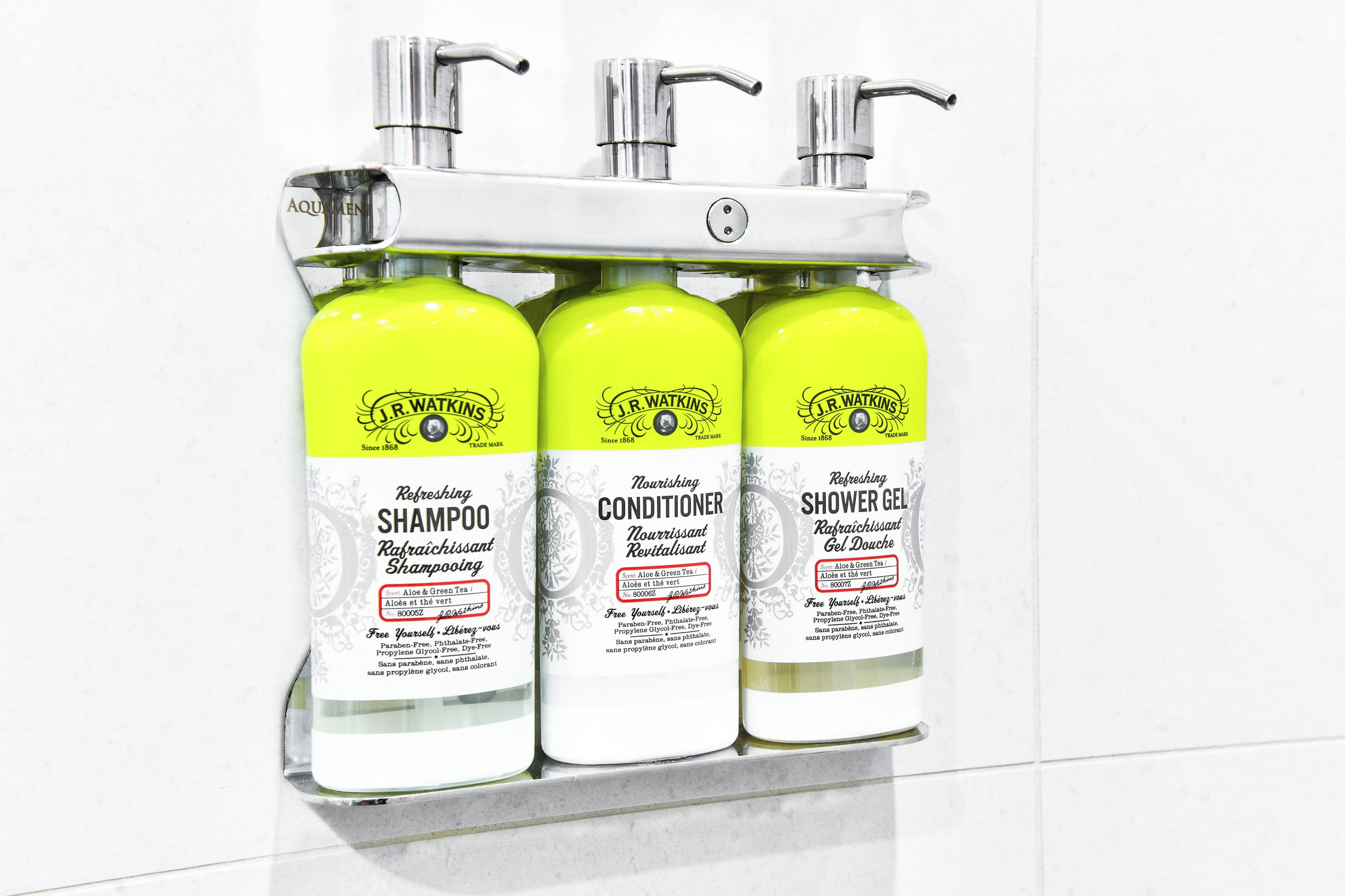 Trois flacons de shampoing, d’après-shampoing et de gel douche de la marque J.R. Watkins, posés sur un mur recouvert de carreaux blancs.