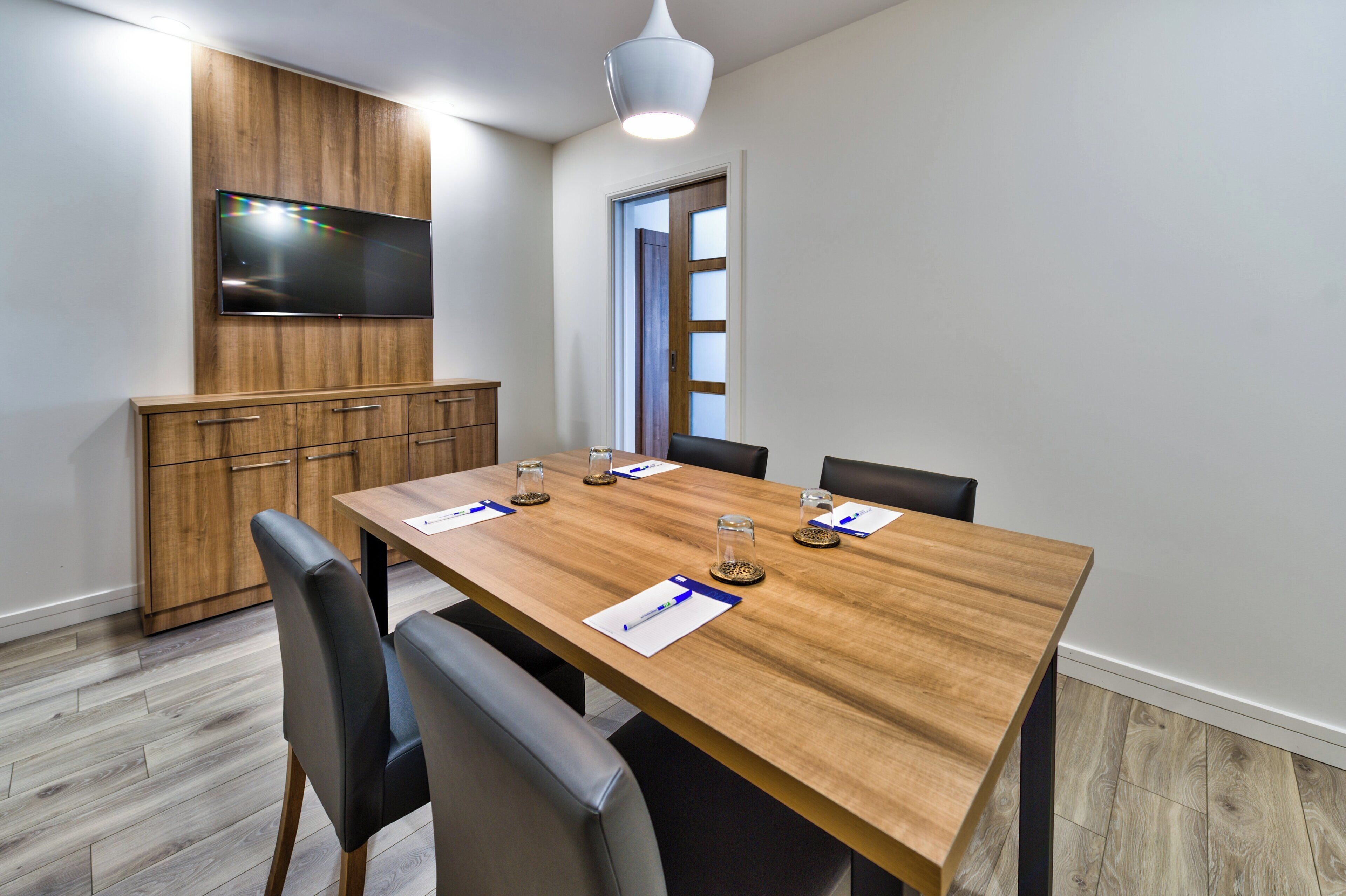 A conference room with a wooden table, four chairs, a television mounted on a wooden panel, and a door with frosted glass panels.