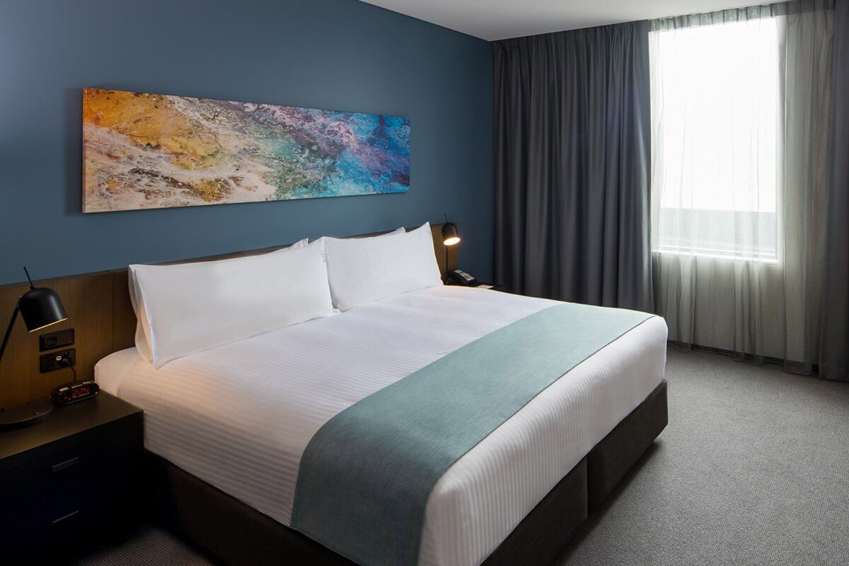 A hotel room with a large bed, a desk, a chair, a painting on the wall, and a window with curtains.