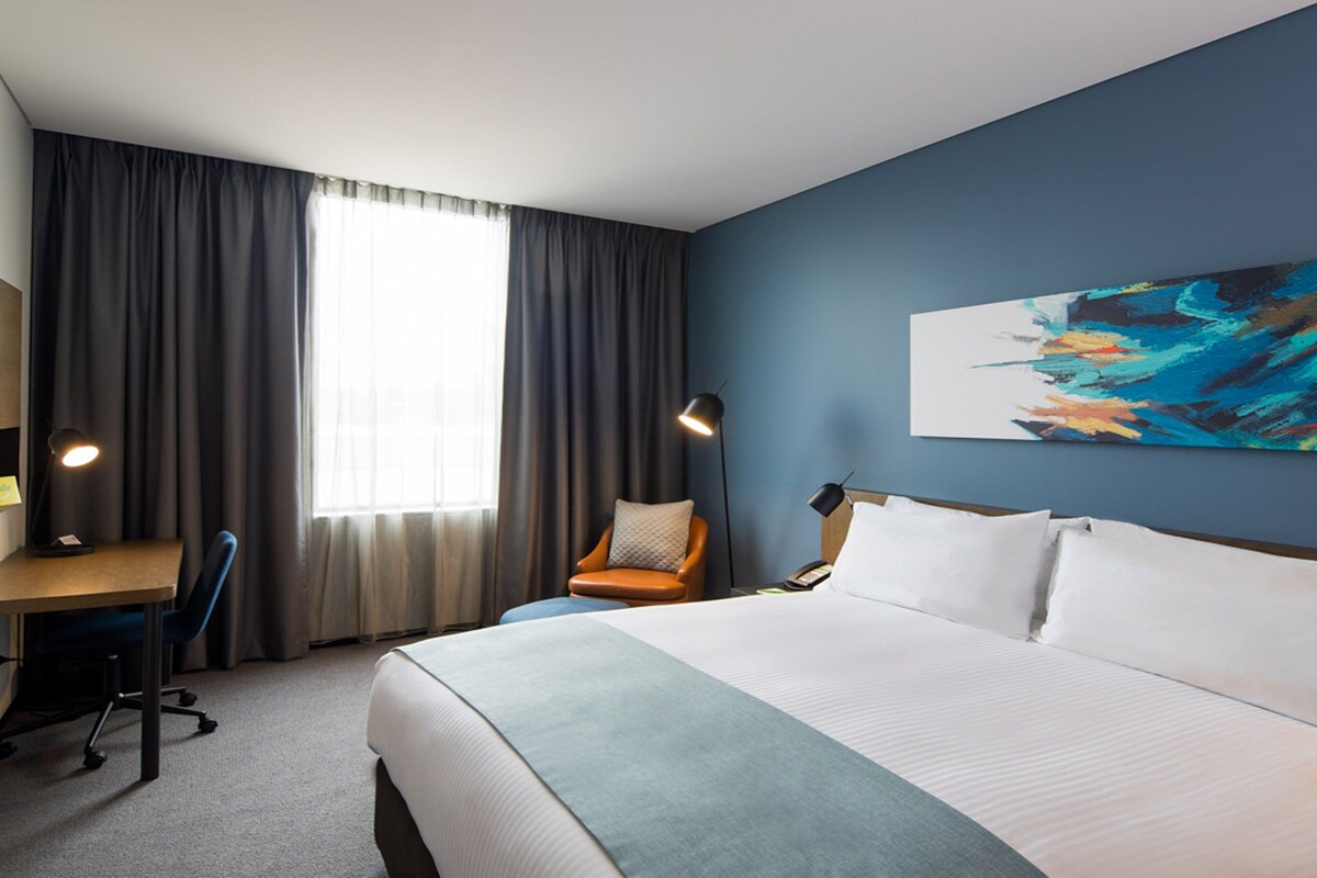 A hotel room with a bed, desk, chair, and a painting on the wall.