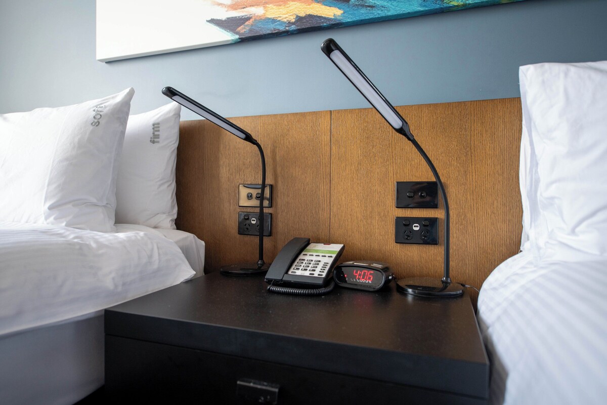 A hotel room with a wooden headboard, a bedside table with a telephone, a digital clock, and two lamps.
