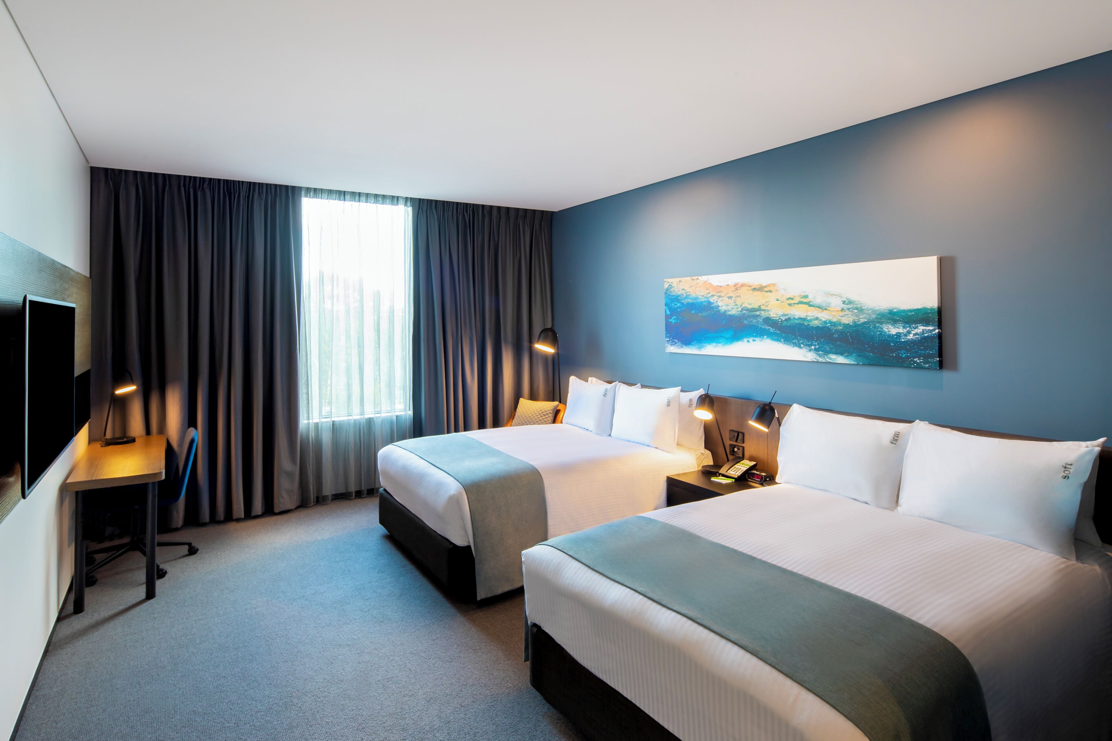 A hotel room with two beds, a desk, a chair, a TV, and a painting on the wall.