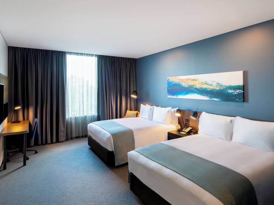 A hotel room with two beds, a desk, a chair, a TV, and a painting on the wall.