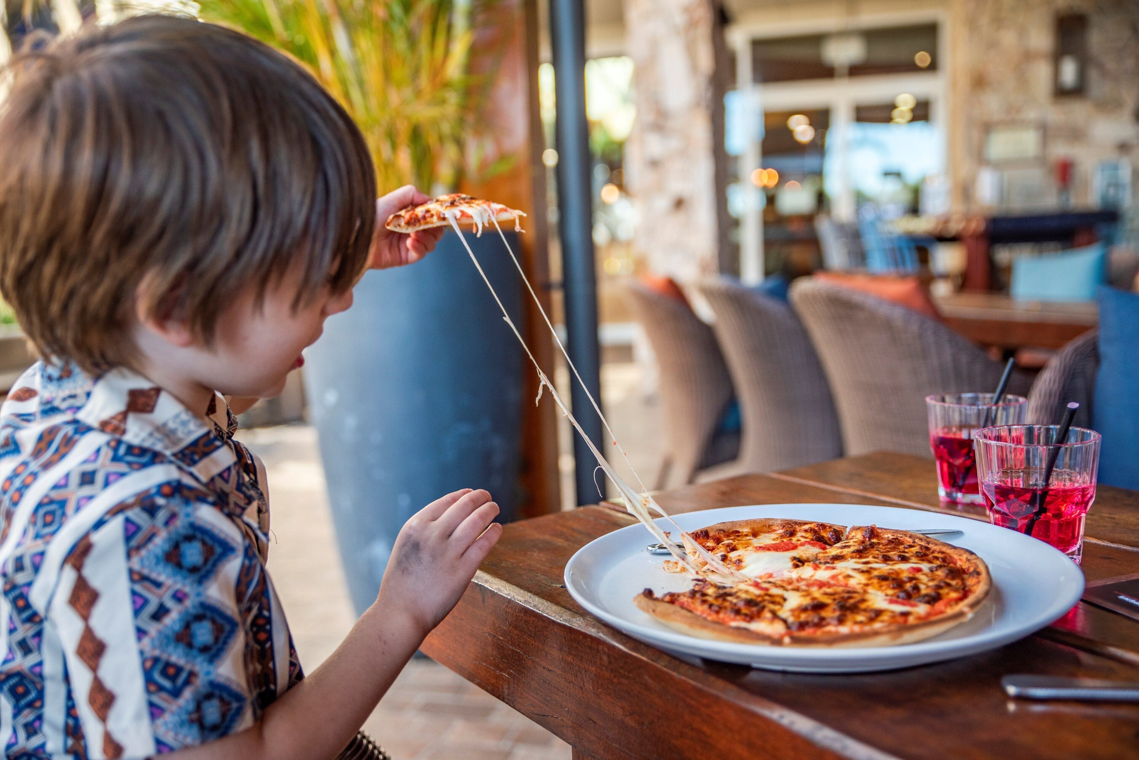 A child holding a slice of pizza with cheese stretching, sitting at an outdoor table with a plate of pizza and two glasses of a red beverage.