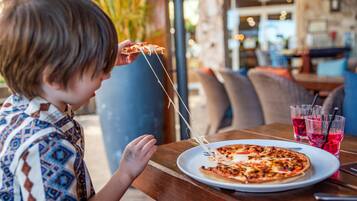 A child holding a slice of pizza with cheese stretching, sitting at an outdoor table with a plate of pizza and two glasses of a red beverage.