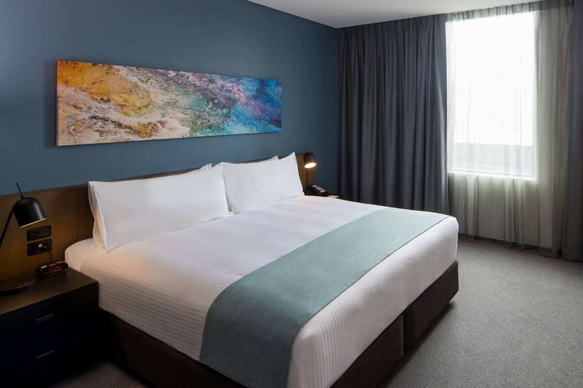 Ein Hotelzimmer mit einem großen Bett, einem Schreibtisch, einem Stuhl, einem Bild an der Wand und einem Fenster mit Vorhängen.