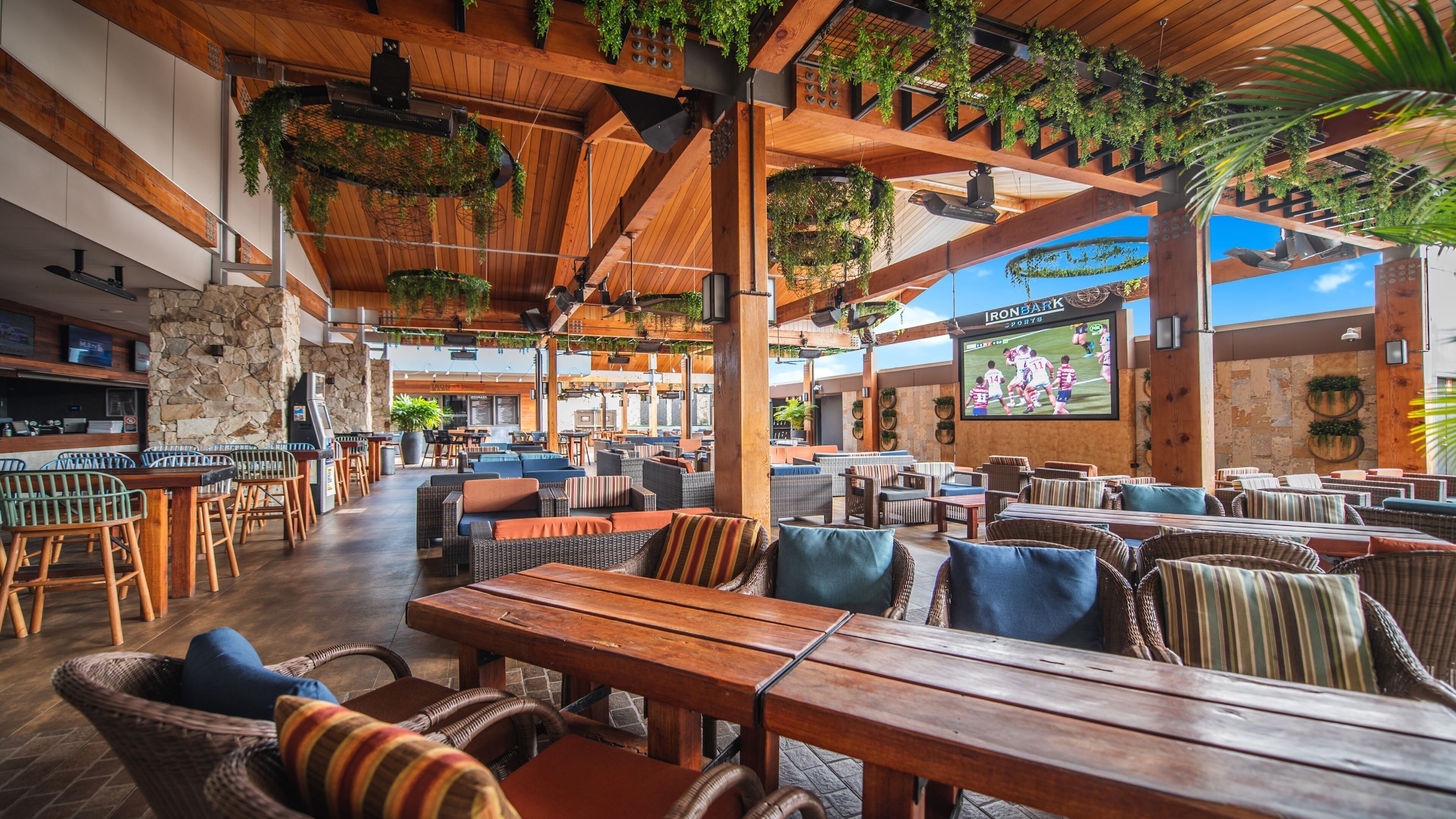 An outdoor dining area with wooden tables and wicker chairs, surrounded by stone walls and greenery.