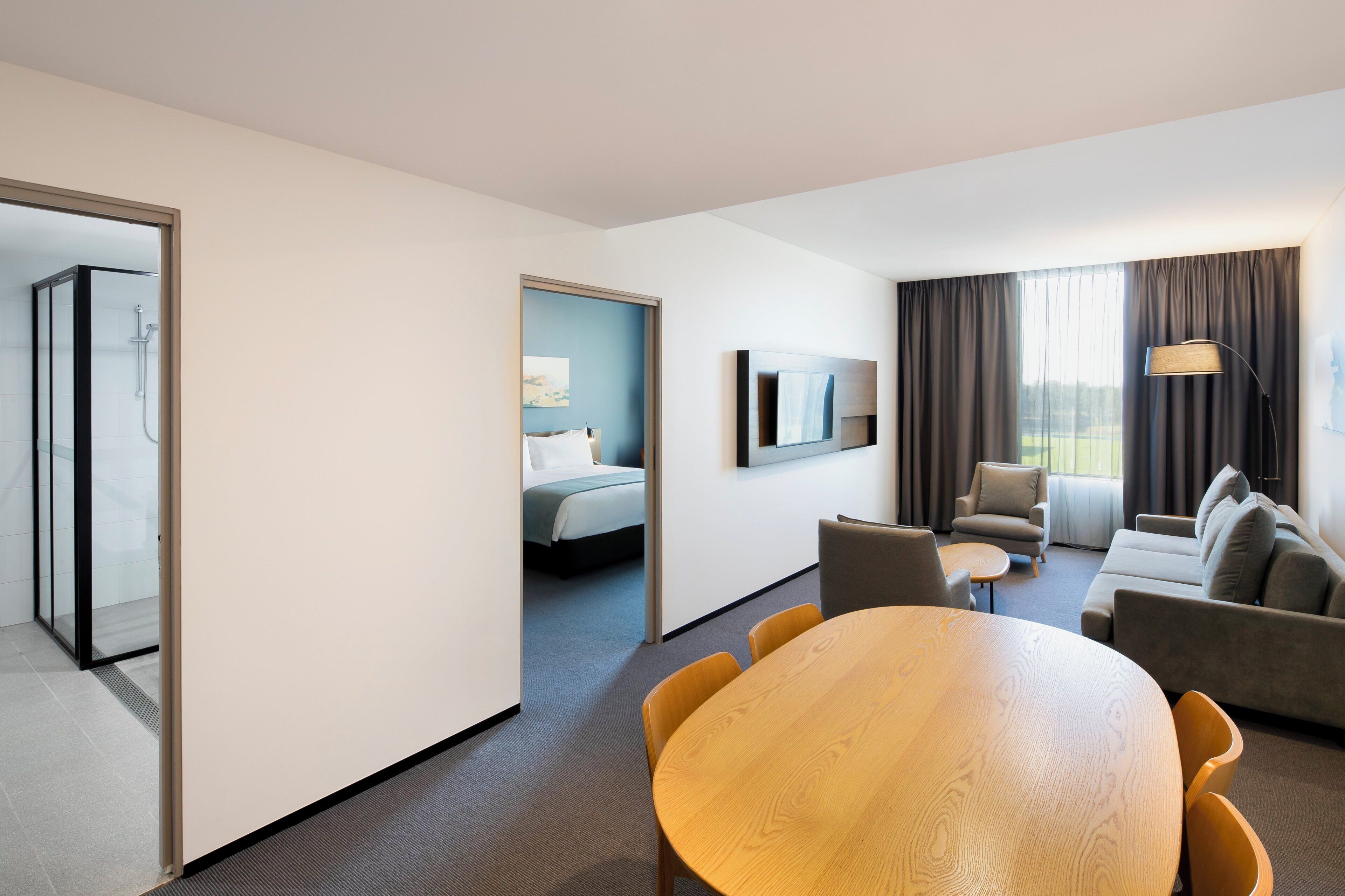A hotel room with a bed, a seating area with a table and chairs, a shower area, and a large window with curtains.
