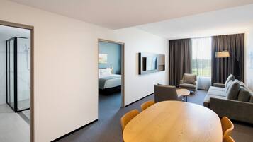 A hotel room with a bed, a seating area with a table and chairs, a shower area, and a large window with curtains.