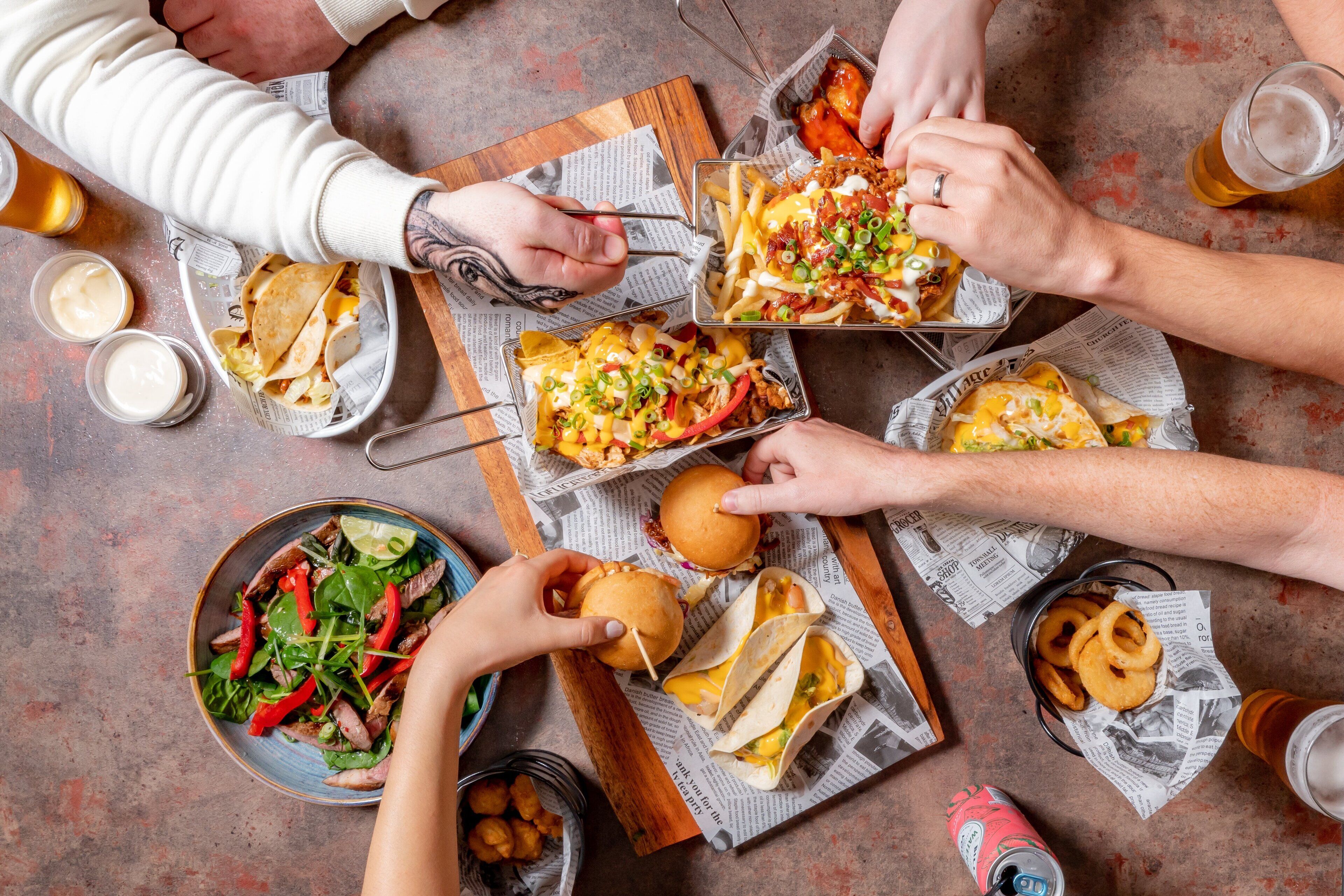 A group of people dining together with various dishes, including tacos, fries, and burgers, on a table with newspapers underneath.