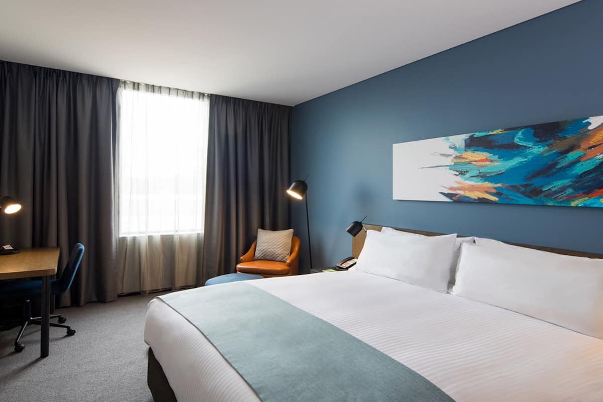 Ein Hotelzimmer mit einem großen Bett, einem Schreibtisch, einem Stuhl, einer Schreibtischlampe, einem Telefon und einem Bild an der Wand.