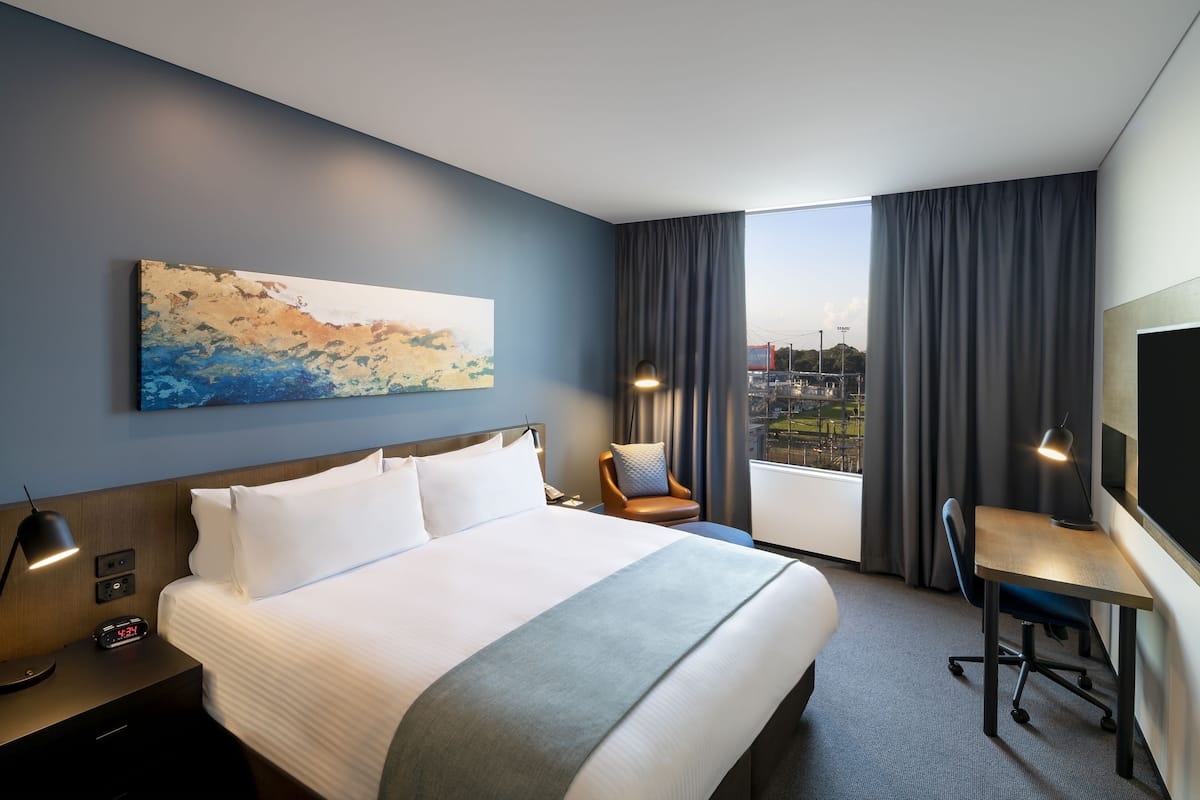 Ein Hotelzimmer mit einem großen Bett, einem Schreibtisch, einem Sessel, einem Bild an der Wand und Blick auf die Stadt durch das Fenster.