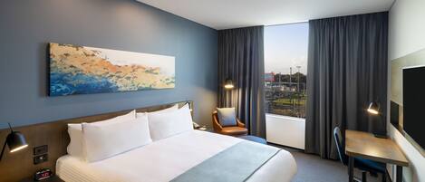 A hotel room with a large bed, a desk, a chair, a painting on the wall, and a view of the city through the window.