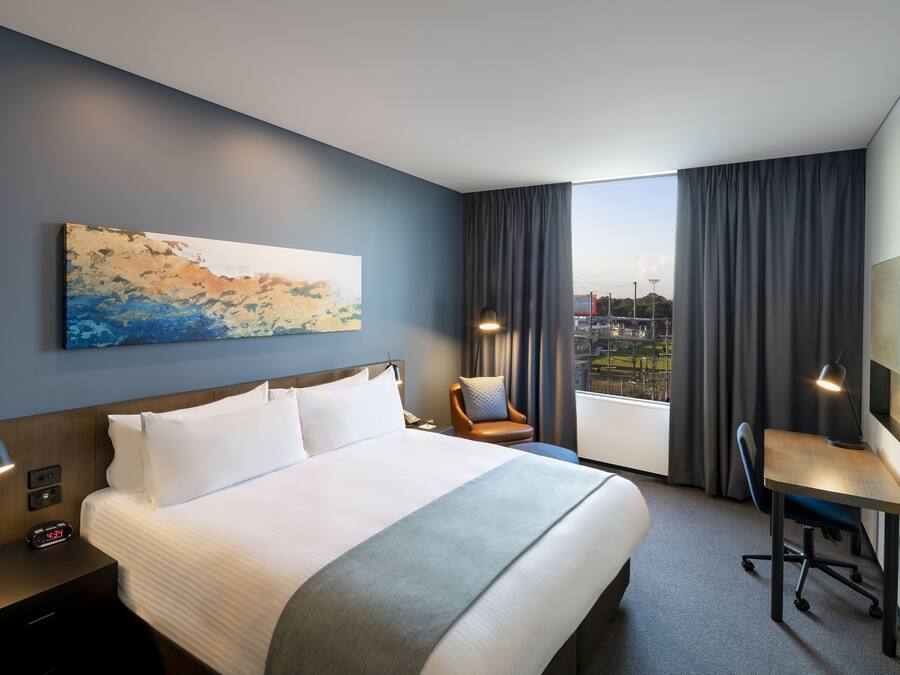 Habitación de hotel con una cama grande, un escritorio, una silla, un cuadro en la pared y vistas de la ciudad a través de la ventana.