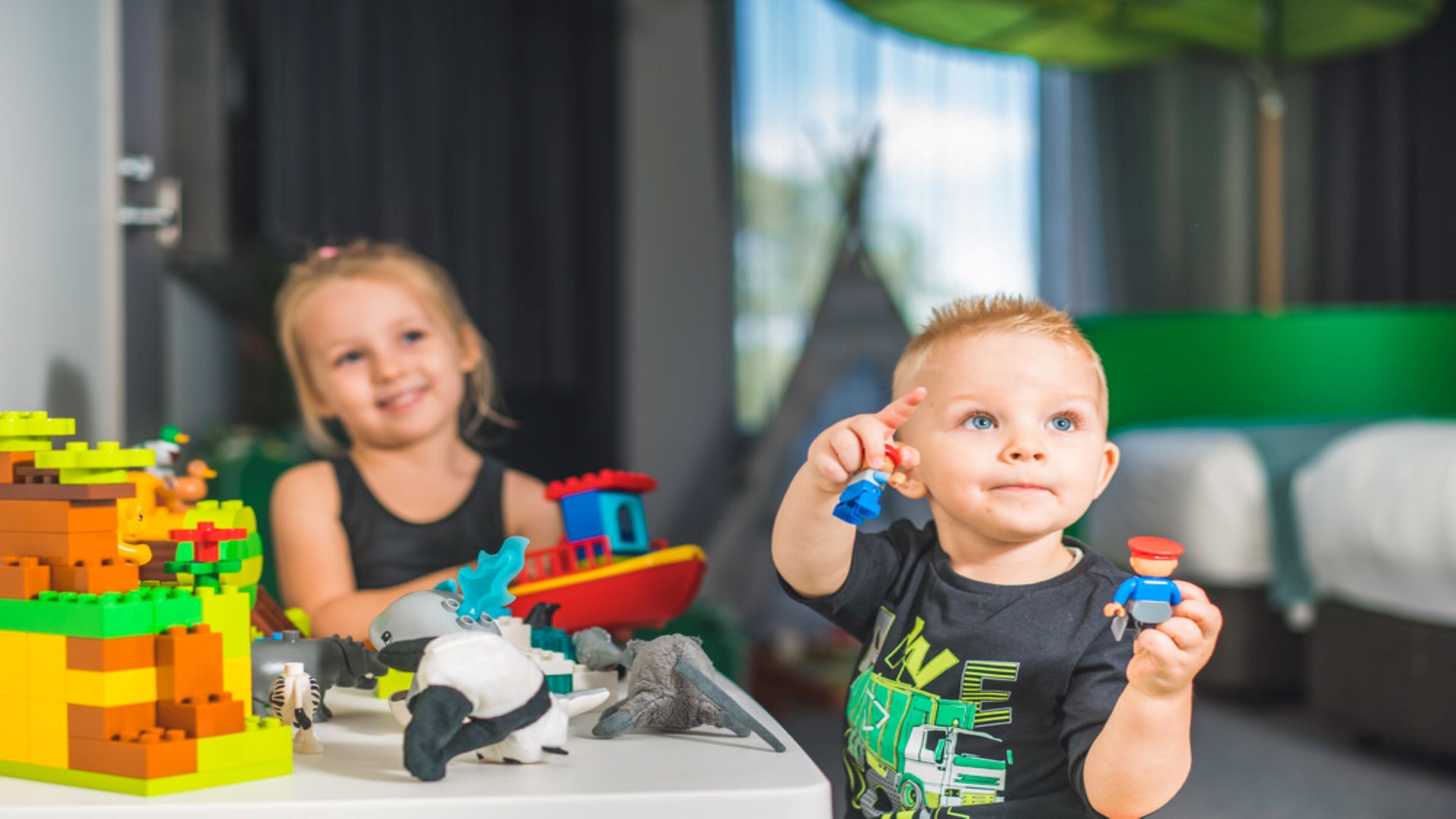 Two children playing with building blocks and toy figures in a room with a bed and a window.