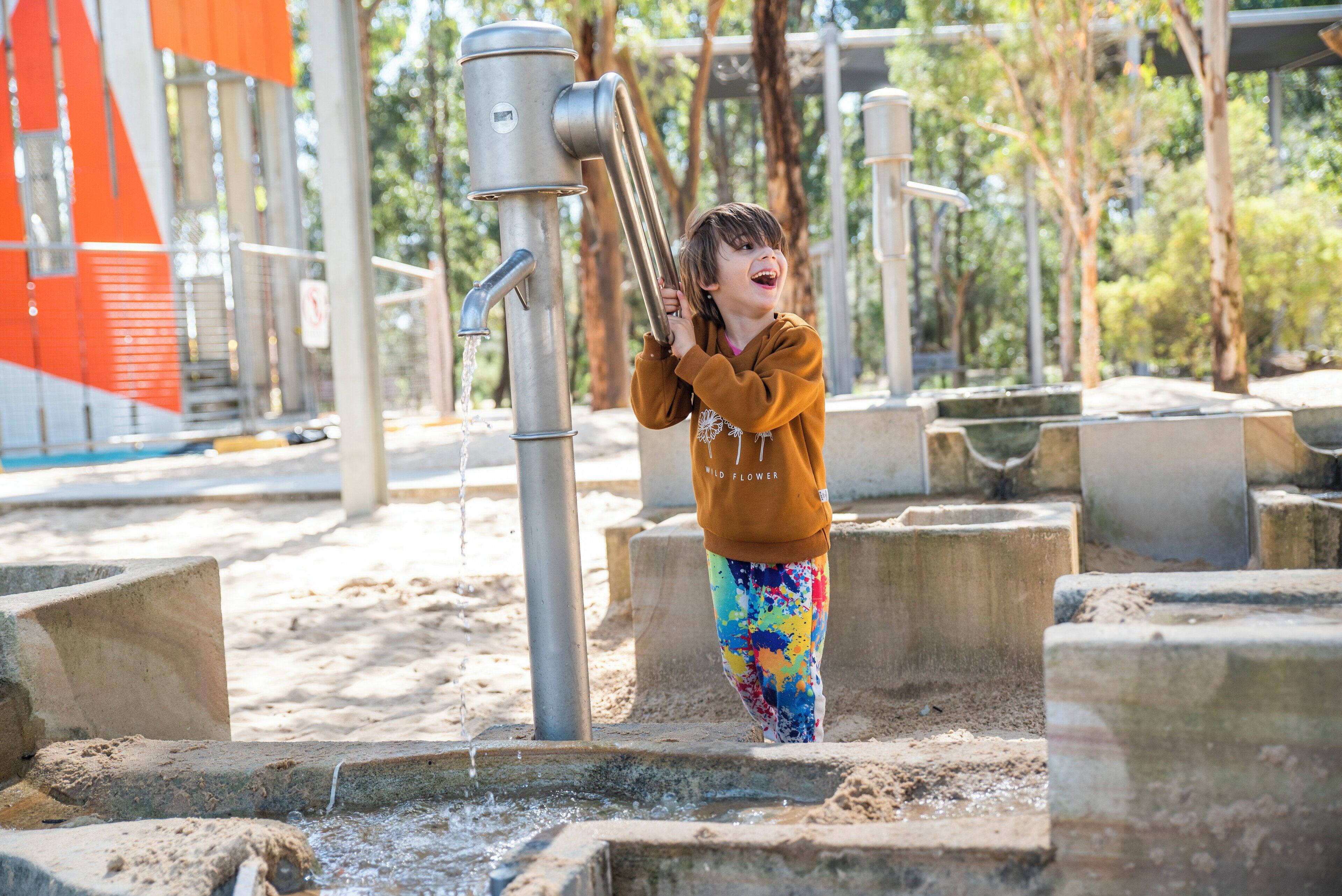 A child playing with a water pump in an outdoor area with concrete steps and metal structures.