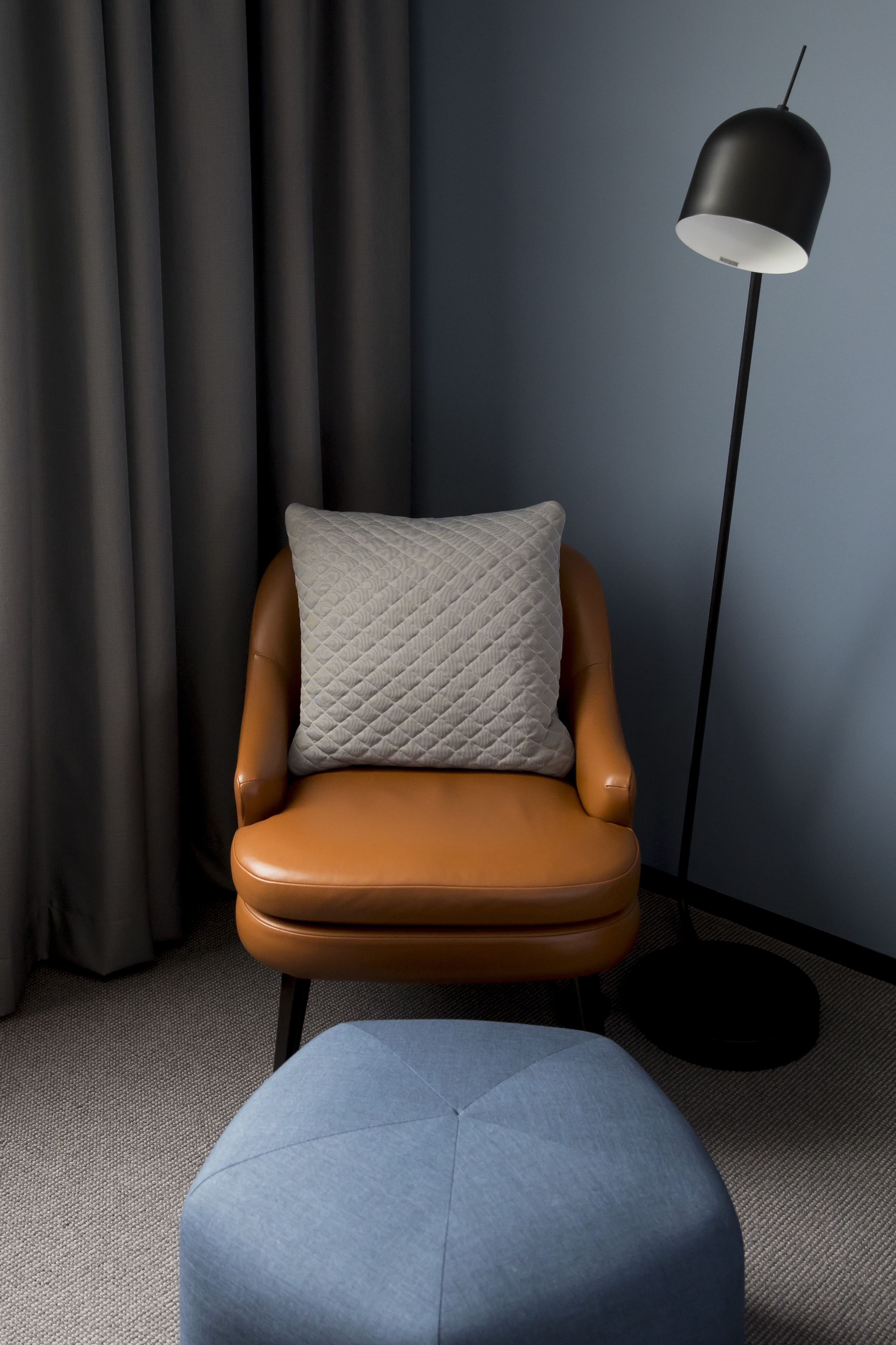 Una habitación moderna con un sillón de cuero marrón, un reposapiés azul, una lámpara de pie negra y cortinas grises.