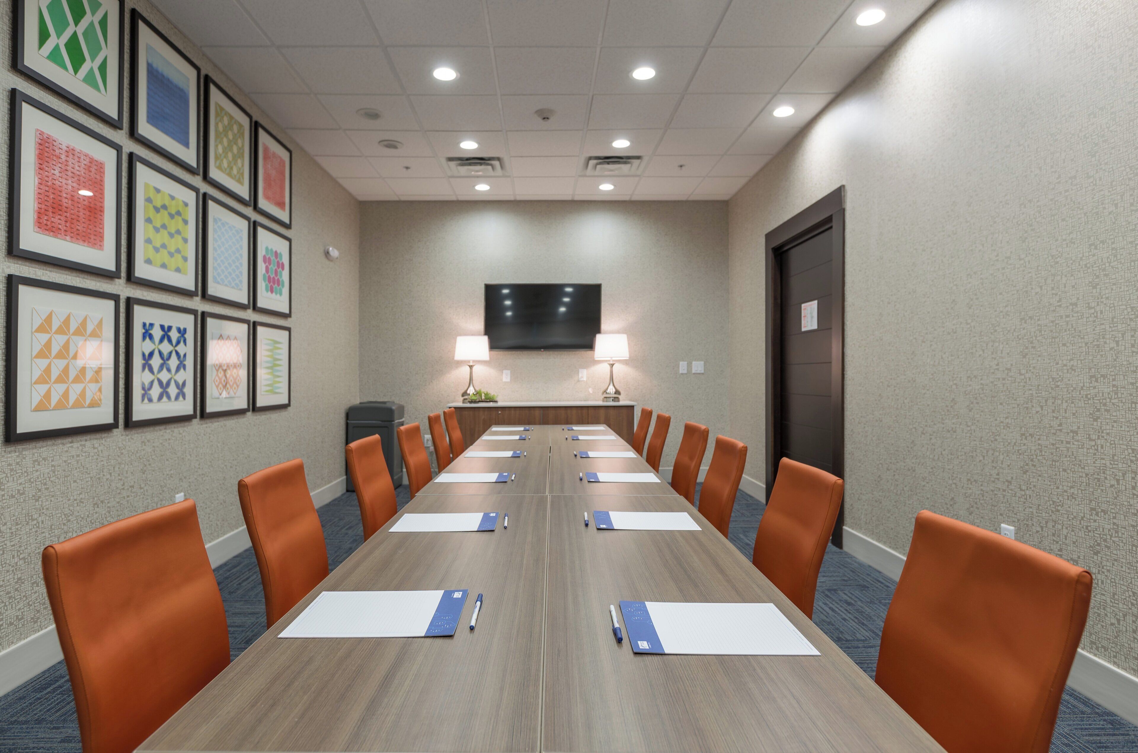 A conference room with a long table, orange chairs, framed artwork on the walls, and a flat-screen TV mounted on the wall.