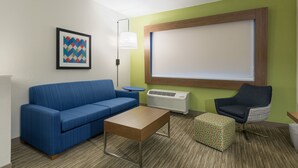 A modern room with a blue sofa, a wooden coffee table, a patterned ottoman, a black armchair, a floor lamp, and a large window with blinds. - Holiday Inn Express and Suites San Marcos South by IHG (San Marcos)
