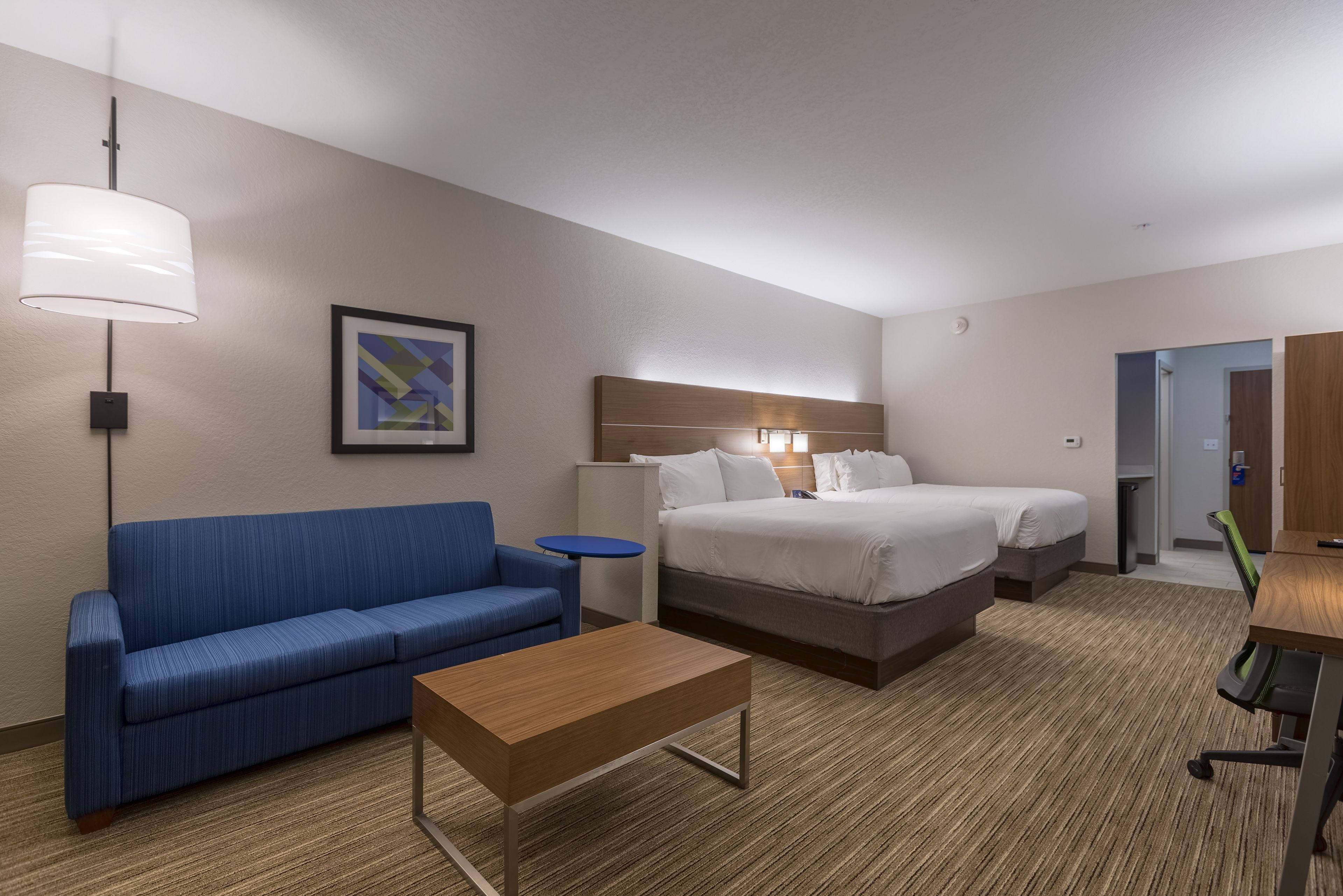 A hotel room with a blue sofa, a wooden coffee table, a bed with white bedding, a bedside table, and a lamp.