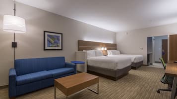 A hotel room with a blue sofa, a wooden coffee table, a bed with white bedding, a bedside table, and a lamp.
