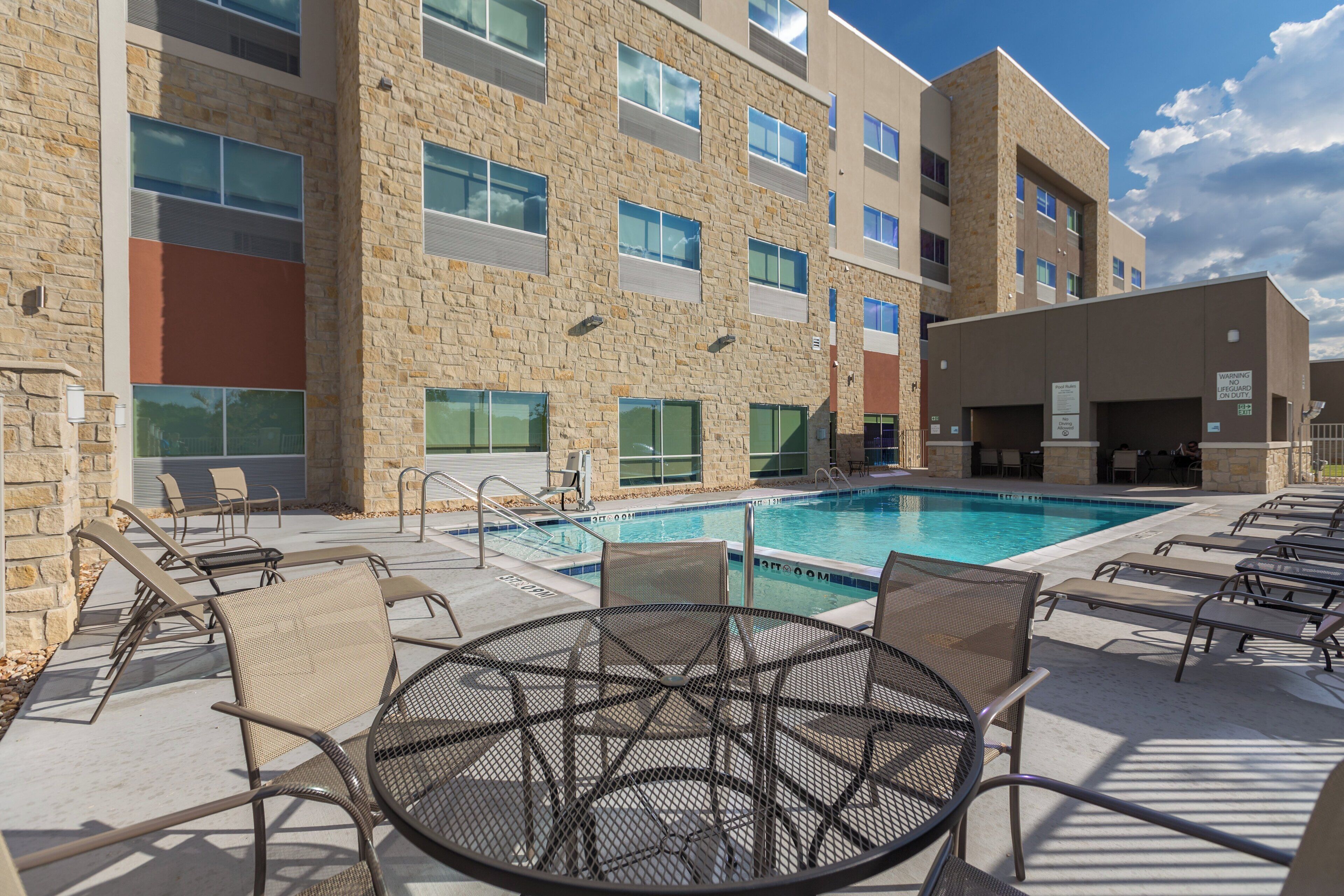Zona de piscina con sillas de descanso y una mesa, rodeada por un edificio de fachada de piedra y varias ventanas.