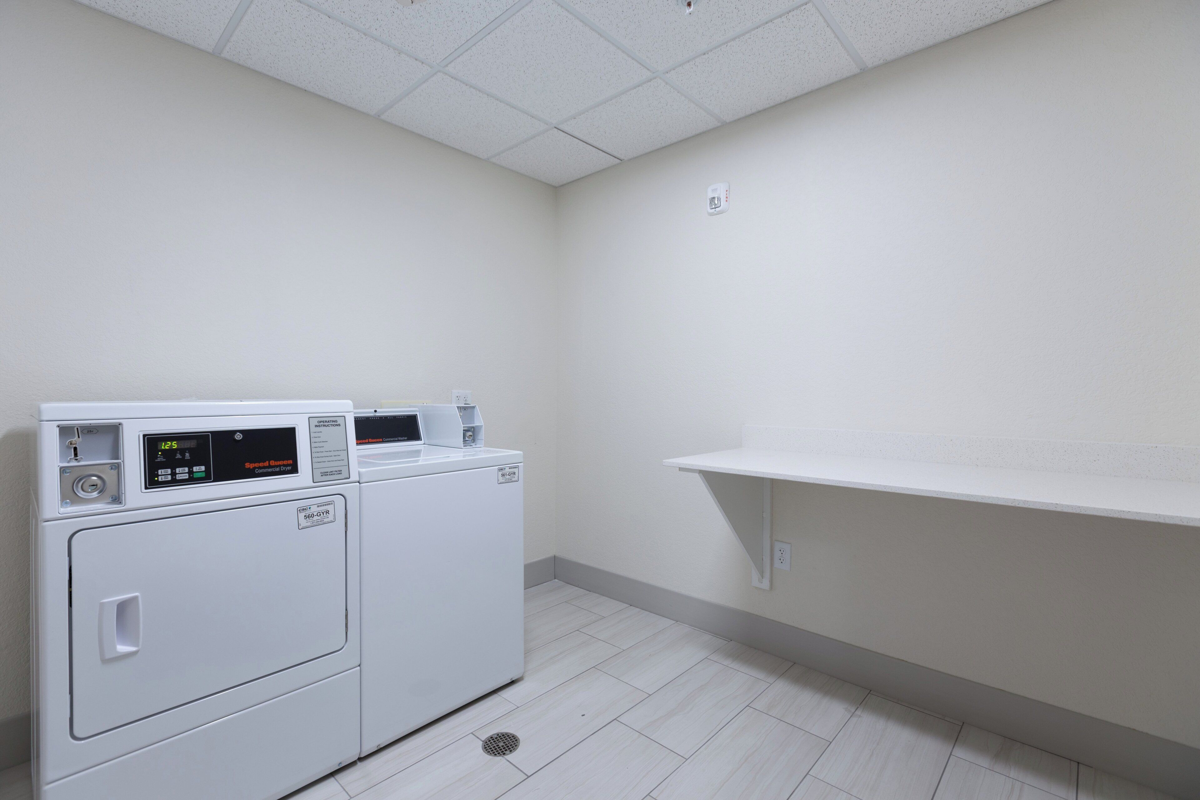 A laundry room with two commercial dryers, a white shelf, and a vent.