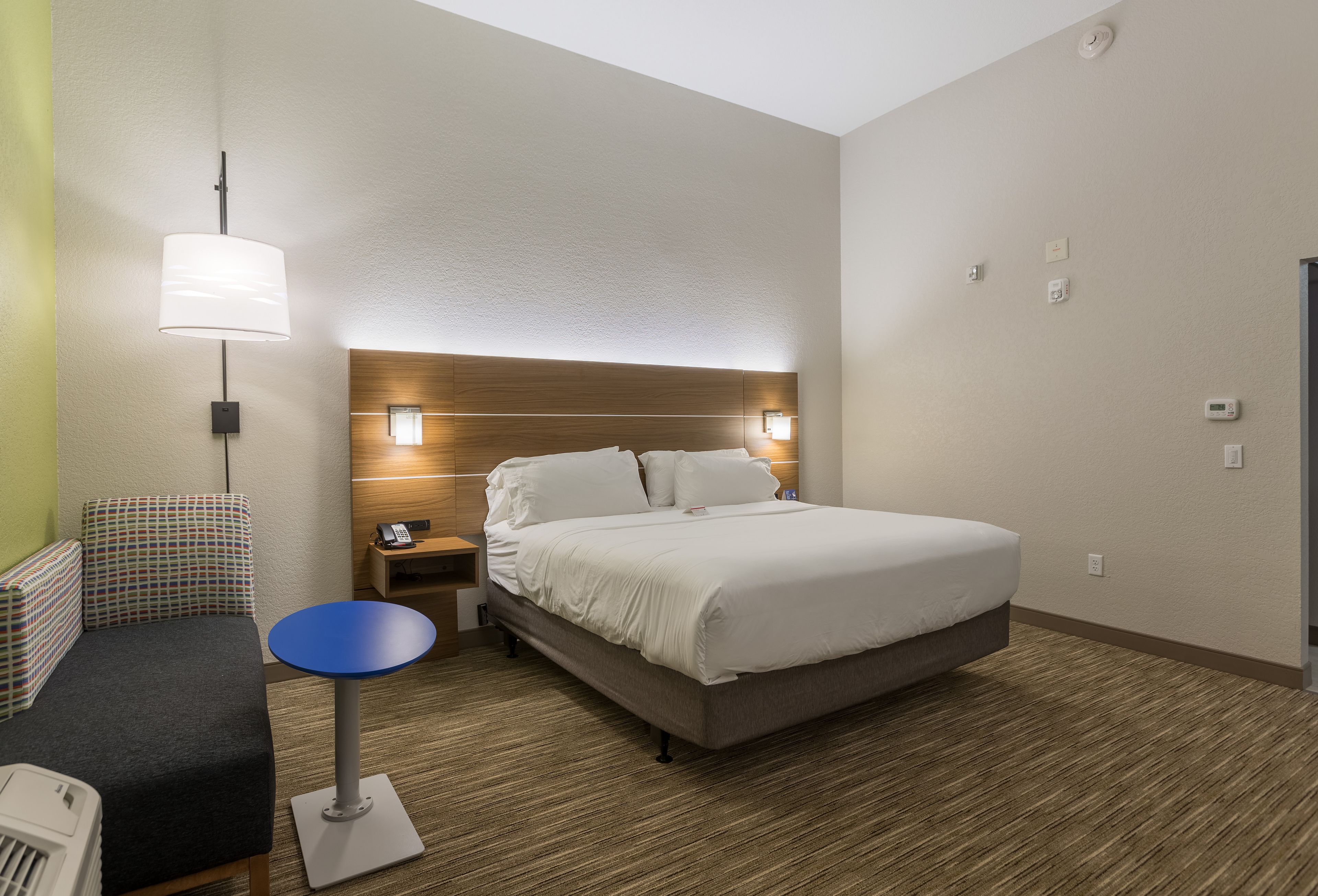Habitación de hotel con cama, mesita de noche, lámpara y una pequeña mesa redonda.