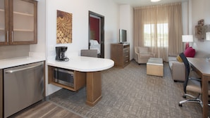 Una habitación de hotel moderna con kitchenette, zona de comedor, sala de estar con sofá y sillón, y un amplio ventanal con cortinas.