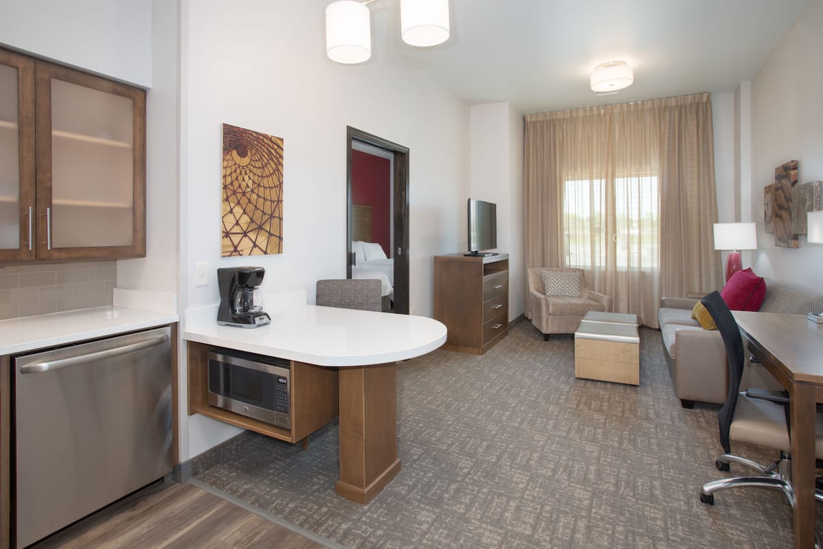 Una habitación de hotel moderna con kitchenette, zona de comedor, sala de estar con sofá y sillón, y un amplio ventanal con cortinas.