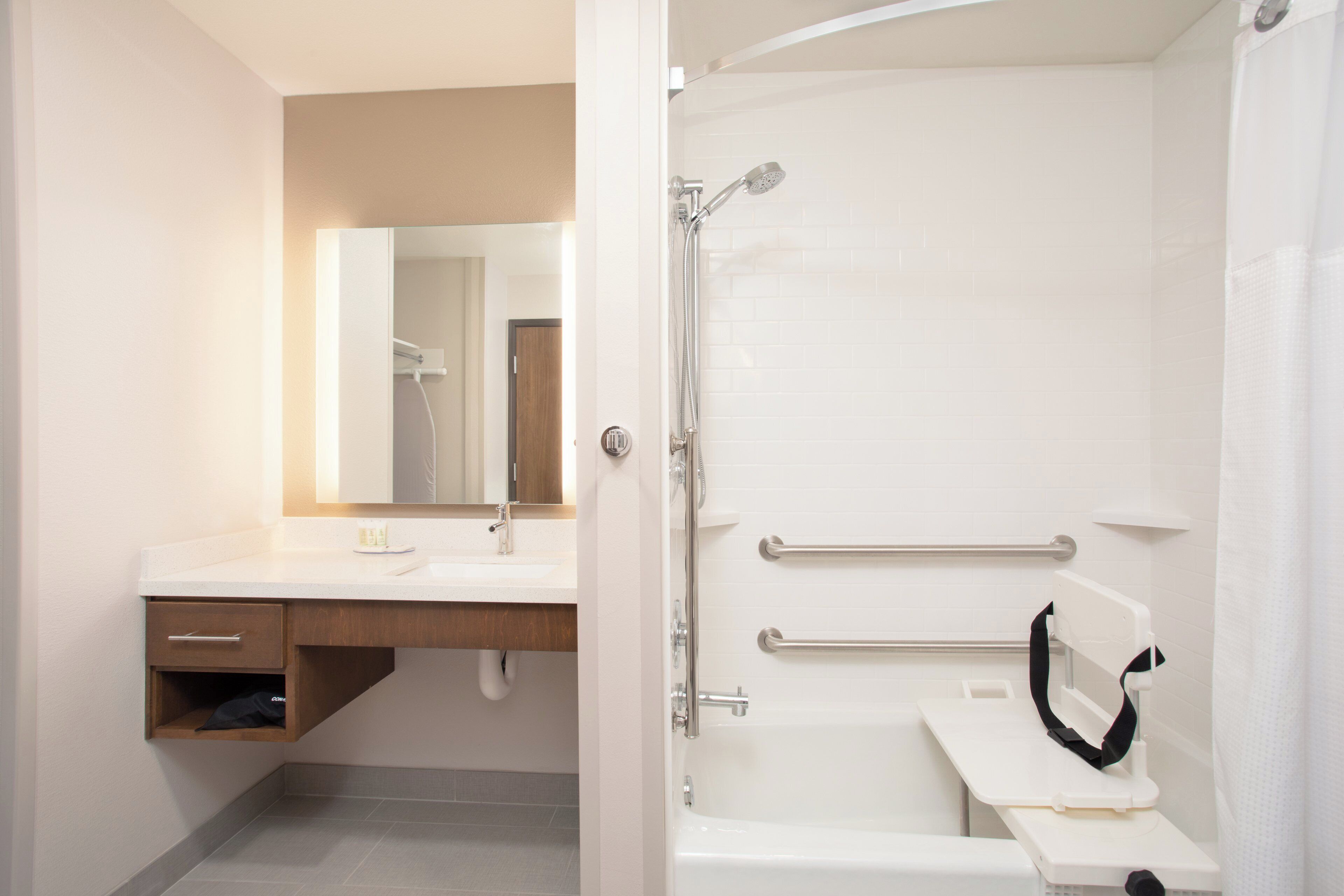 Een badkamer met een witte wastafel, spiegel en douche. Er zijn handgrepen in de douche en een handdoekhouder.