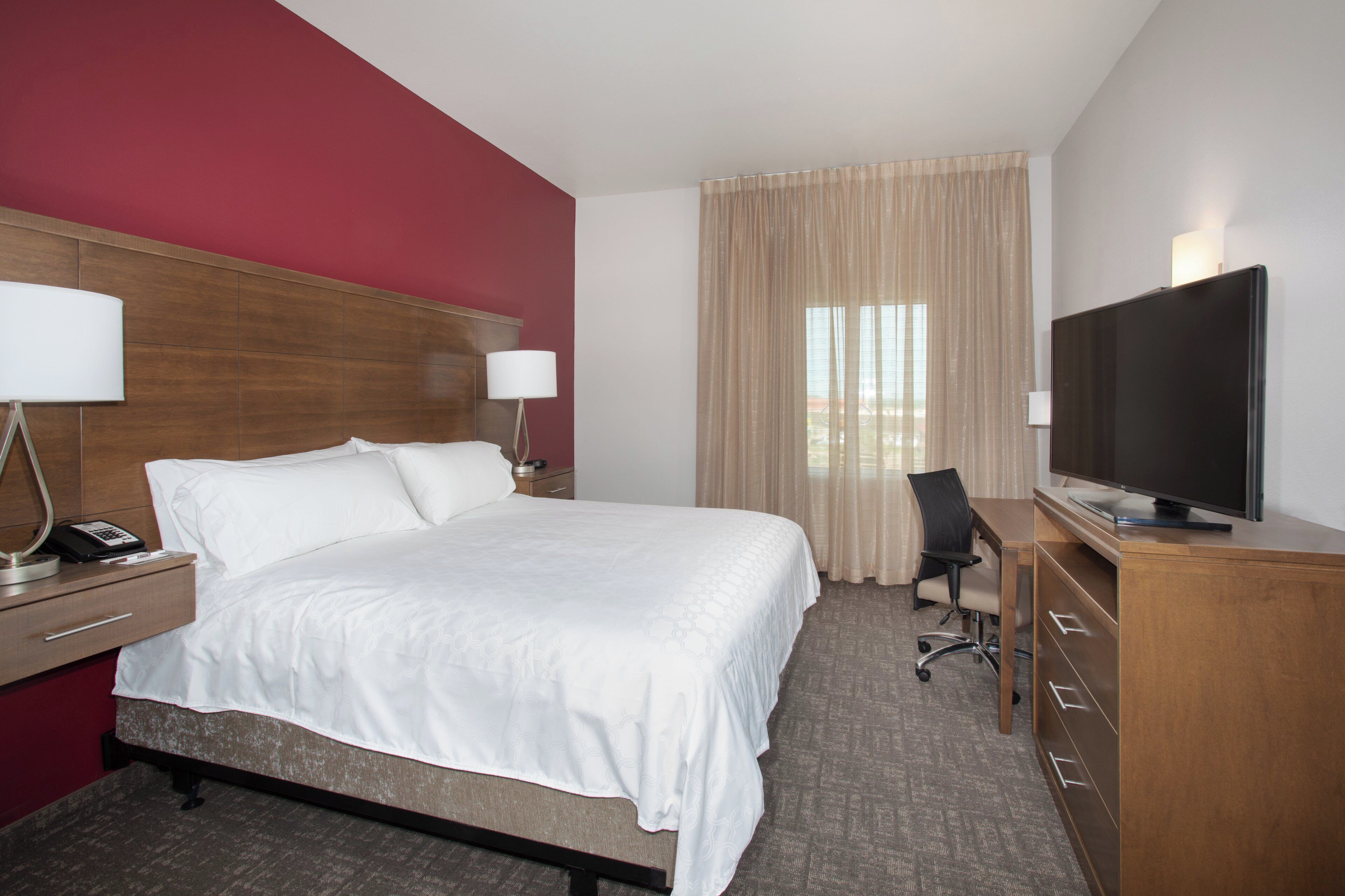 A hotel room with a bed, a desk with a lamp, a chair, a television, and a window with curtains.