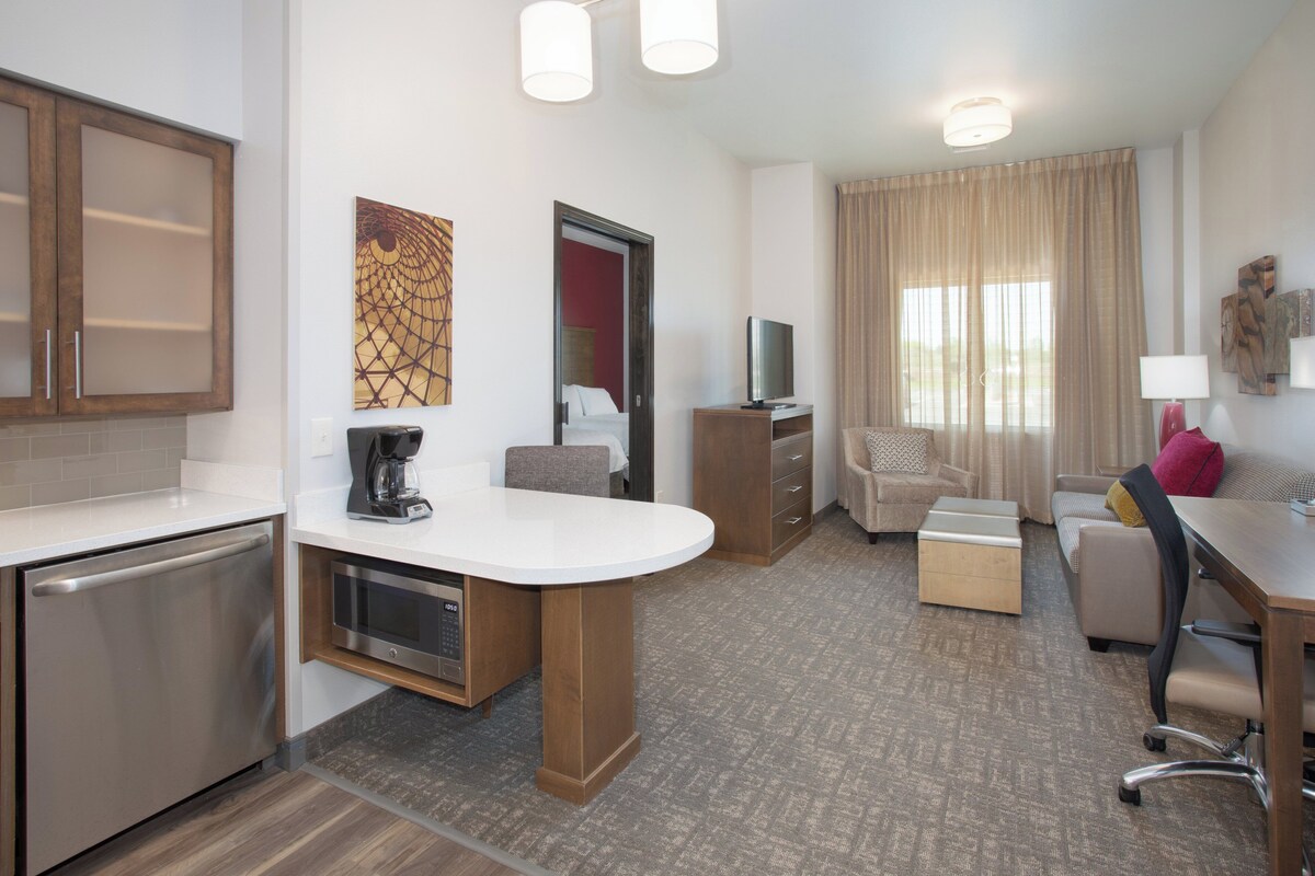 Habitación de hotel con kitchenette, zona de comedor, espacio de descanso con sofá y sillones, y un amplio ventanal con cortinas.