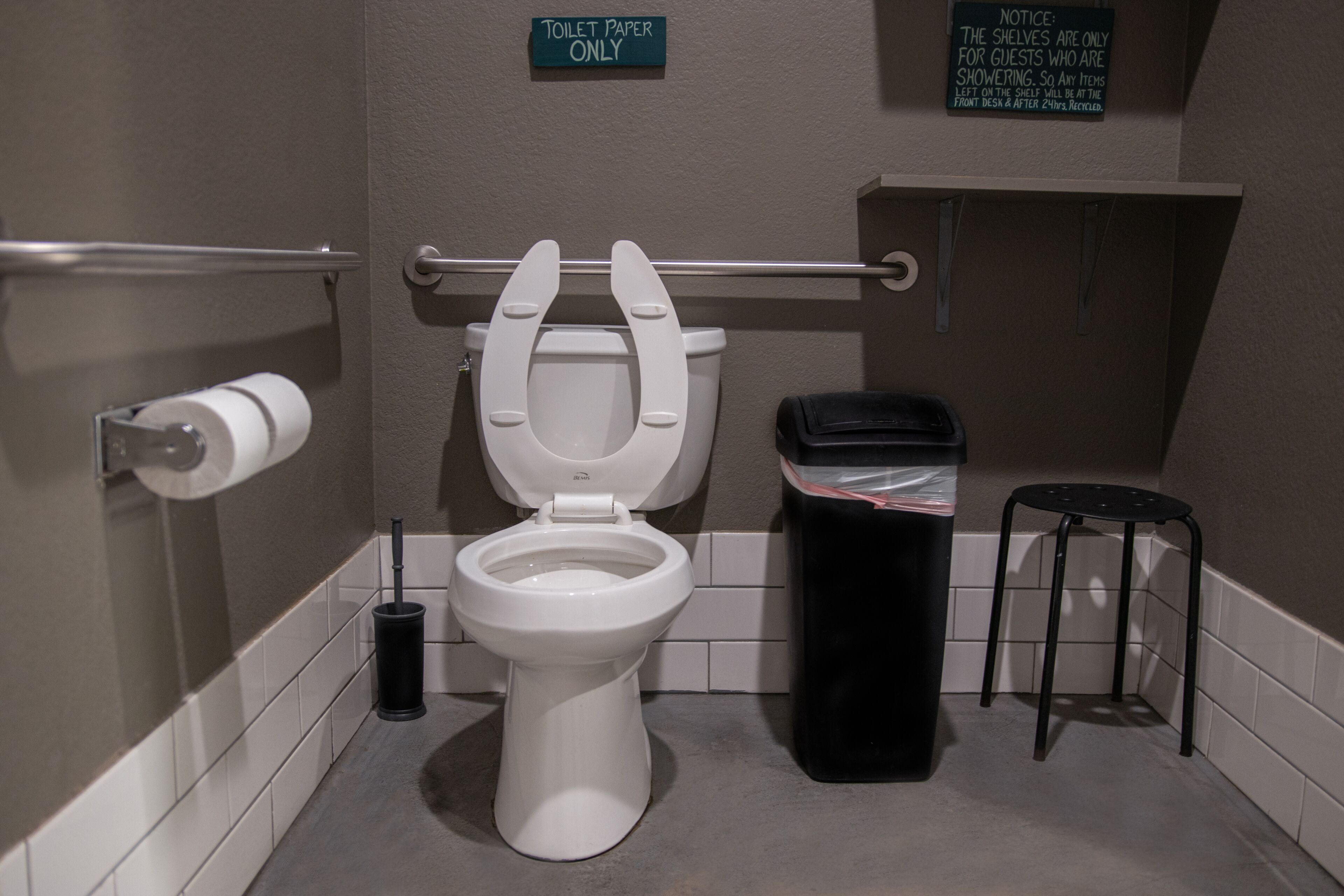 A bathroom with a toilet, a trash bin, and a shelf with a notice.