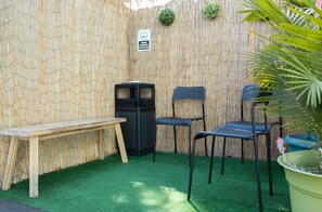 A small outdoor seating area with a wooden bench and two chairs, a trash bin, and a bamboo wall.