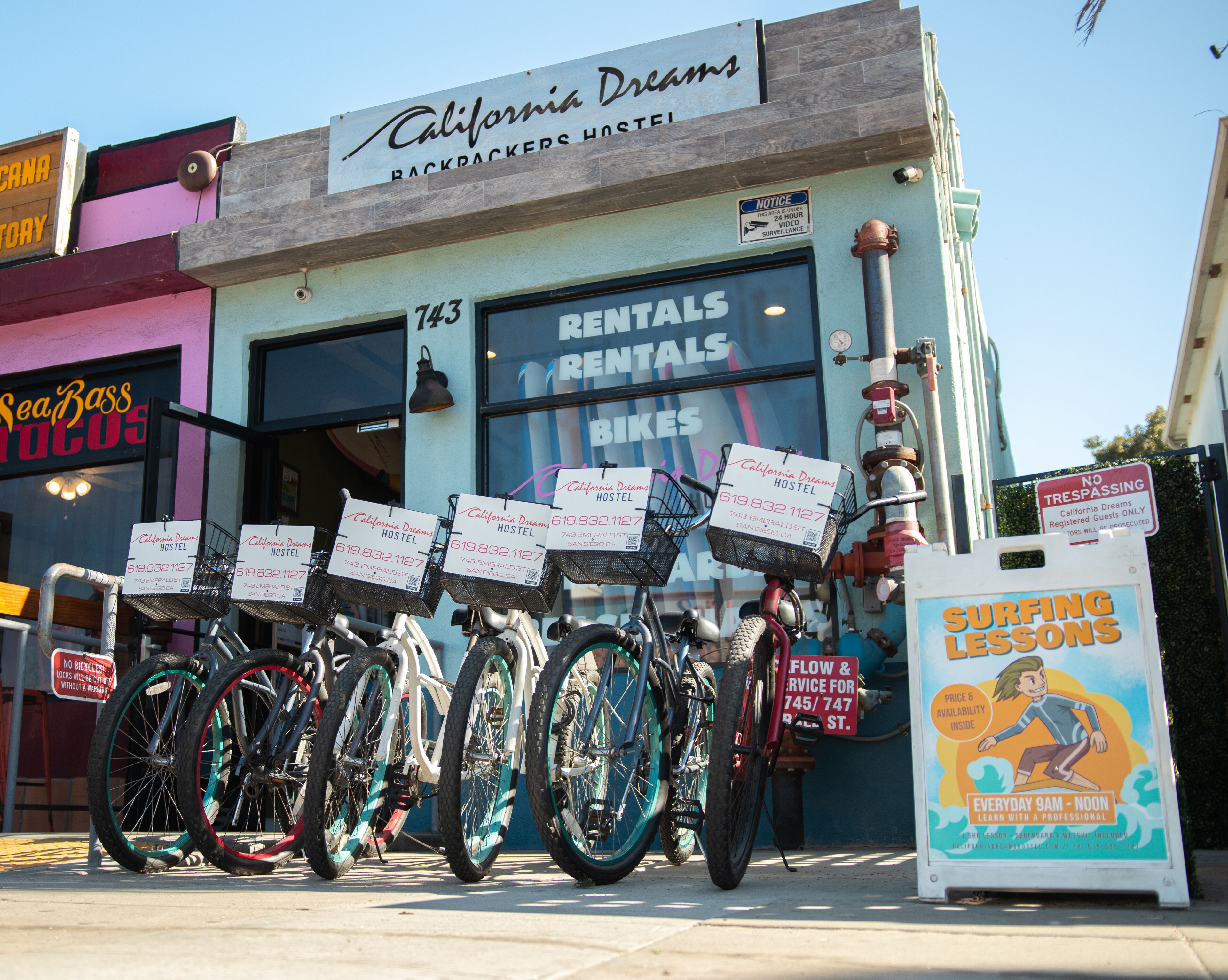 A hostel named California Dreams with a signboard, rental bikes, and a surf lessons advertisement.