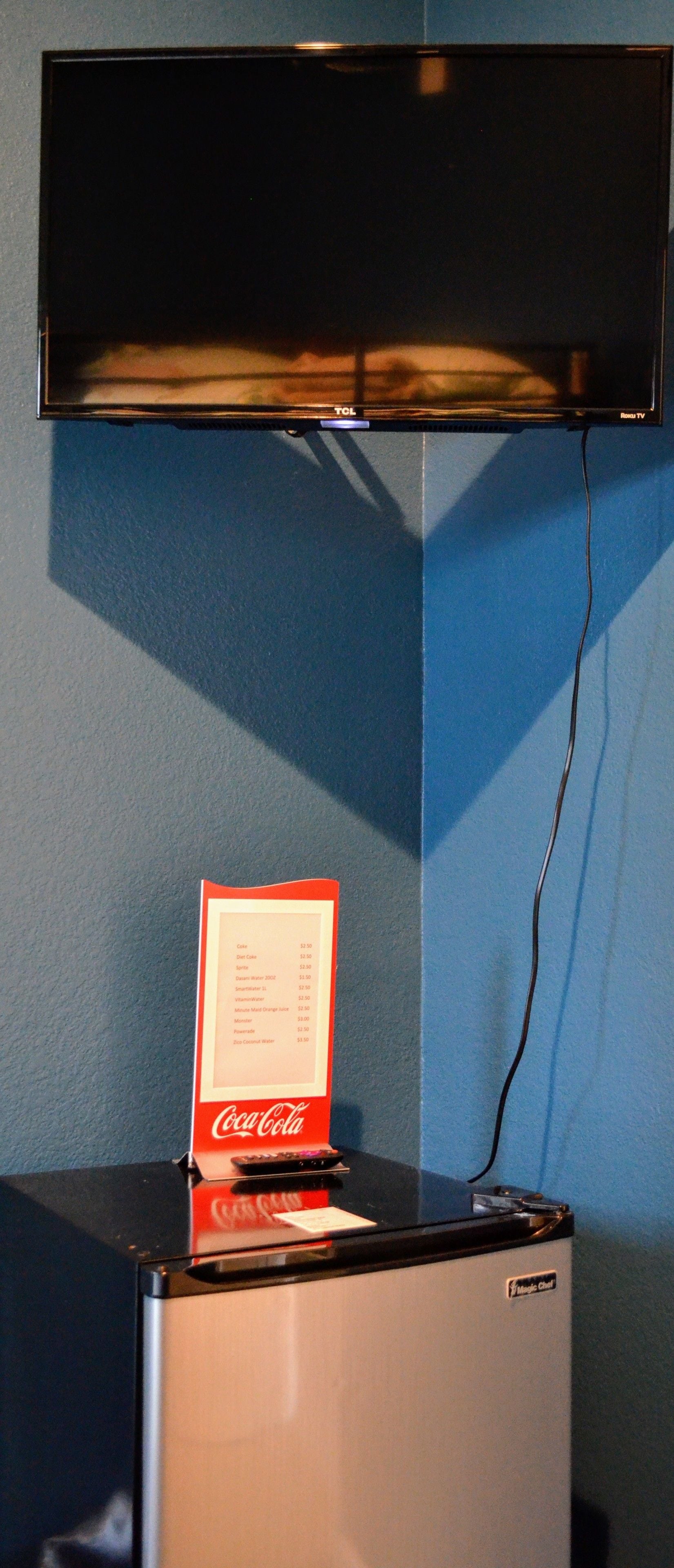 A refrigerator with a Coca-Cola vending machine on top, a black TV above, and a cable running down the wall.