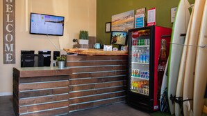 A reception desk with a Coca-Cola refrigerator, surfboards, and a monitor displaying a list. - California Dreams Hostel - Pacific Beach (San Diego)