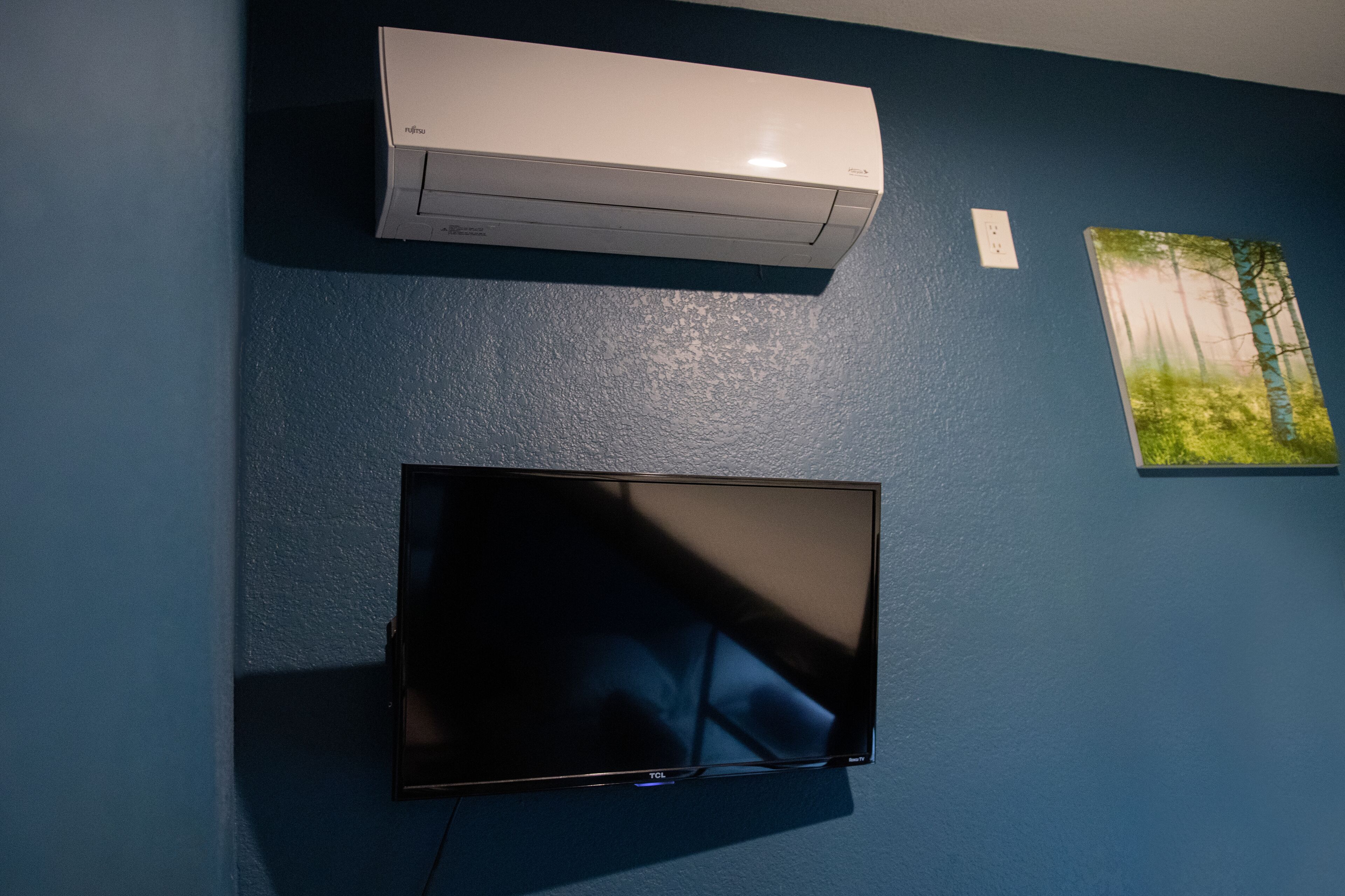 A room with a flat-screen TV mounted on the wall, a white air conditioner above it, and a painting of a forest scene to the right.