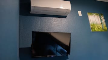 A room with a flat-screen TV mounted on the wall, a white air conditioner above it, and a painting of a forest scene to the right.
