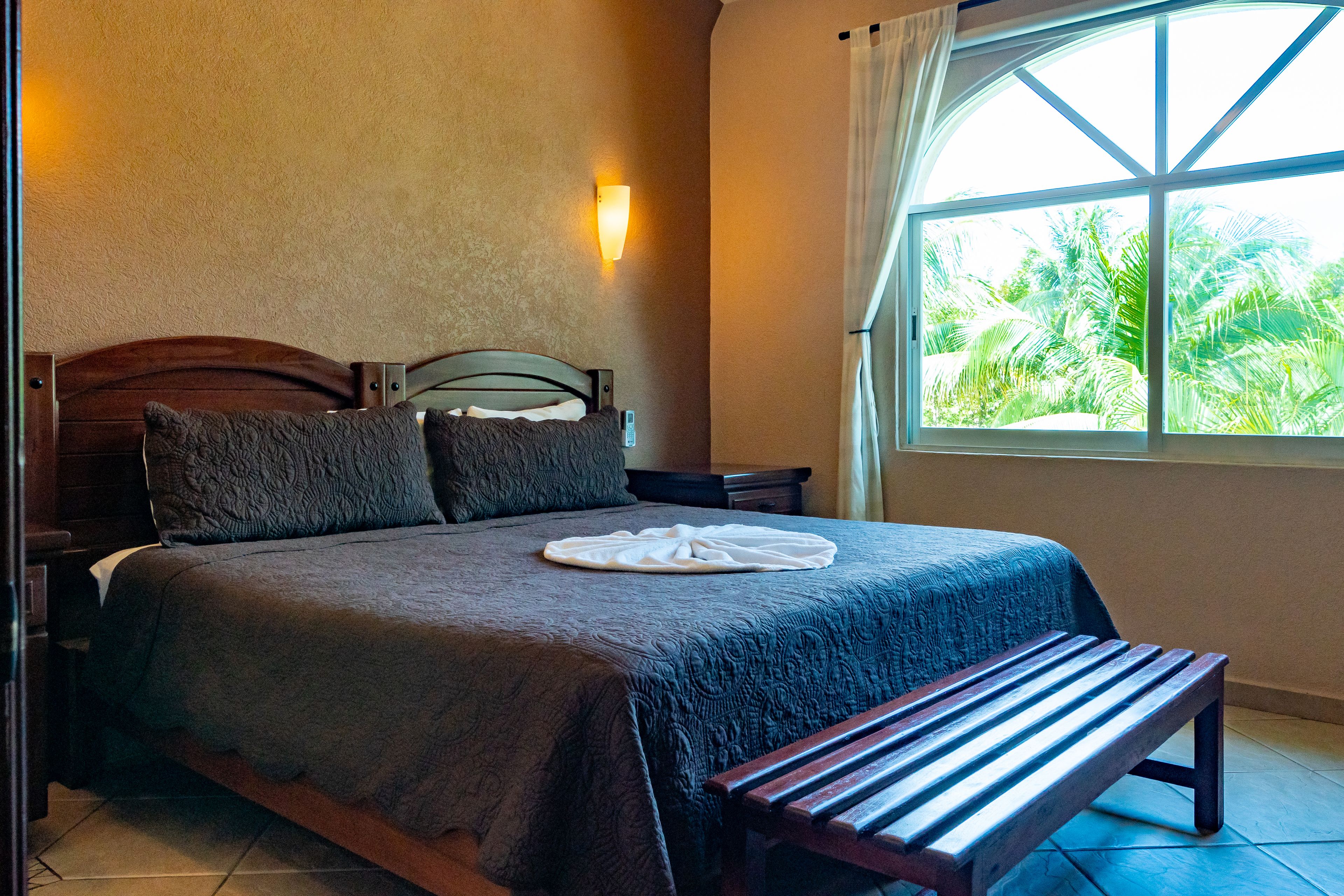 A neatly made bed with a dark bedspread, a wooden bench at the foot of the bed, and a wall-mounted light fixture.